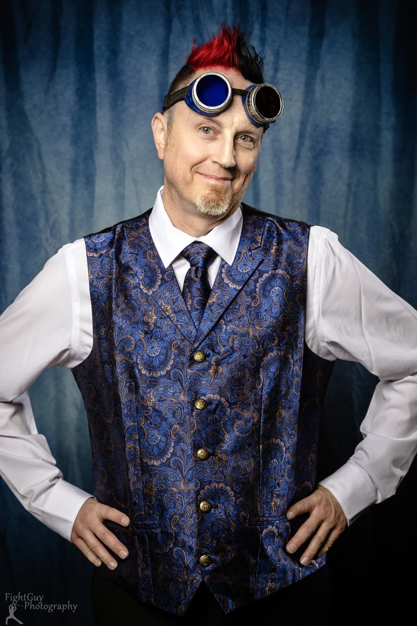 A man with red and black spiked hair wearing goggles, a white shirt, a patterned blue vest, and a tie, standing with hands on hips against a blue background.