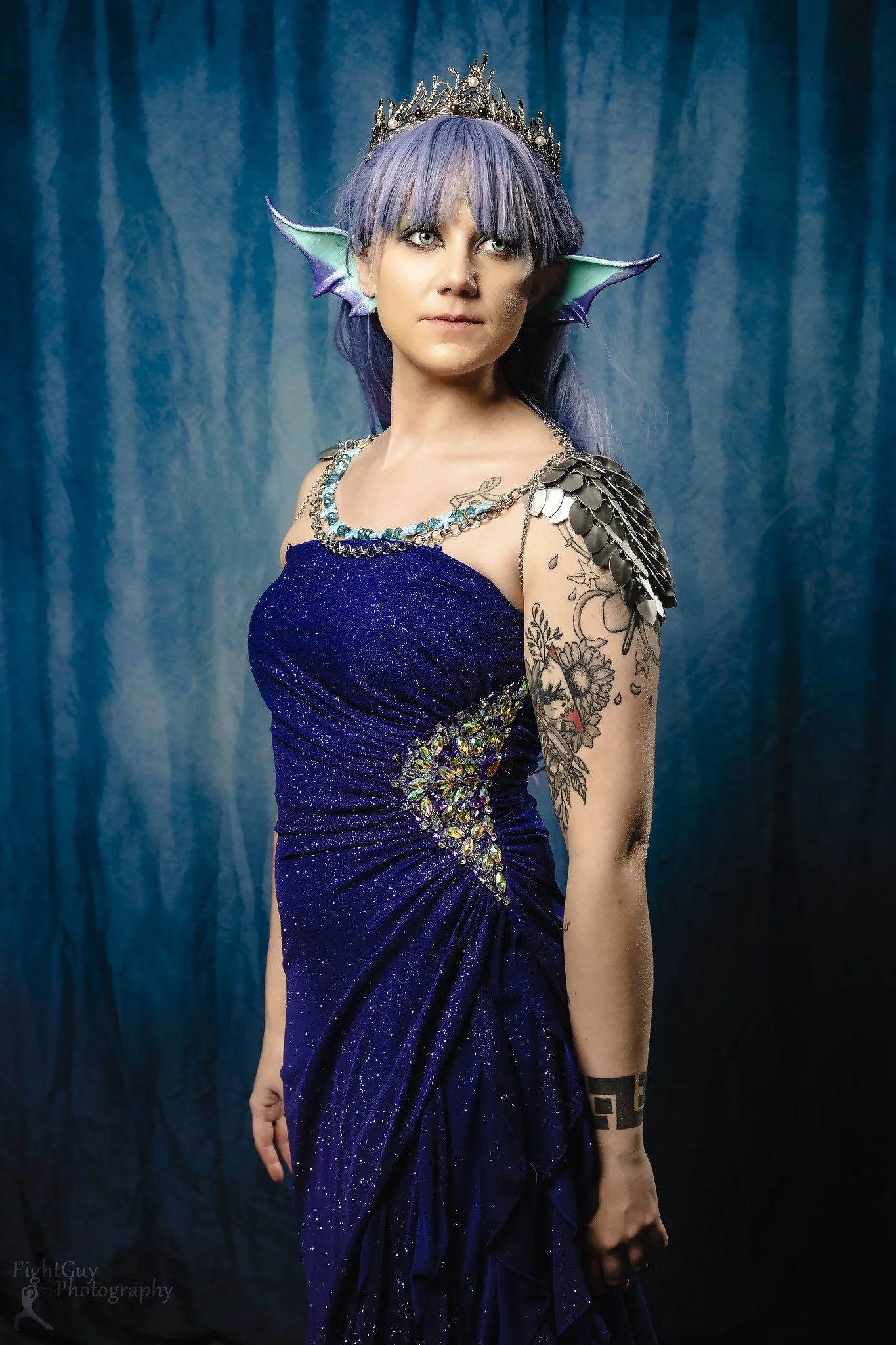 A woman with purple hair and fantasy elf ears wearing a sparkly blue gown, a tiara, and decorative shoulder armor, standing against a blue backdrop.