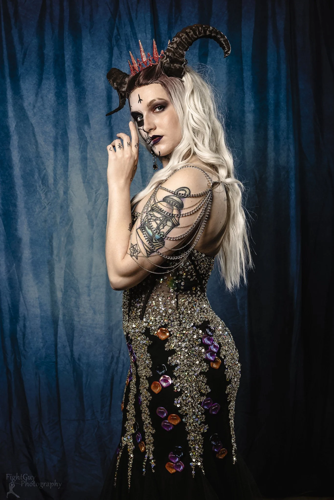 A woman with long, wavy platinum blonde hair dressed in a dark, jewel-embellished dress with silver chains on her shoulder, wearing a headpiece with horns, a red crown, and a skull face paint with black lipstick, against a blue curtain backdrop.
