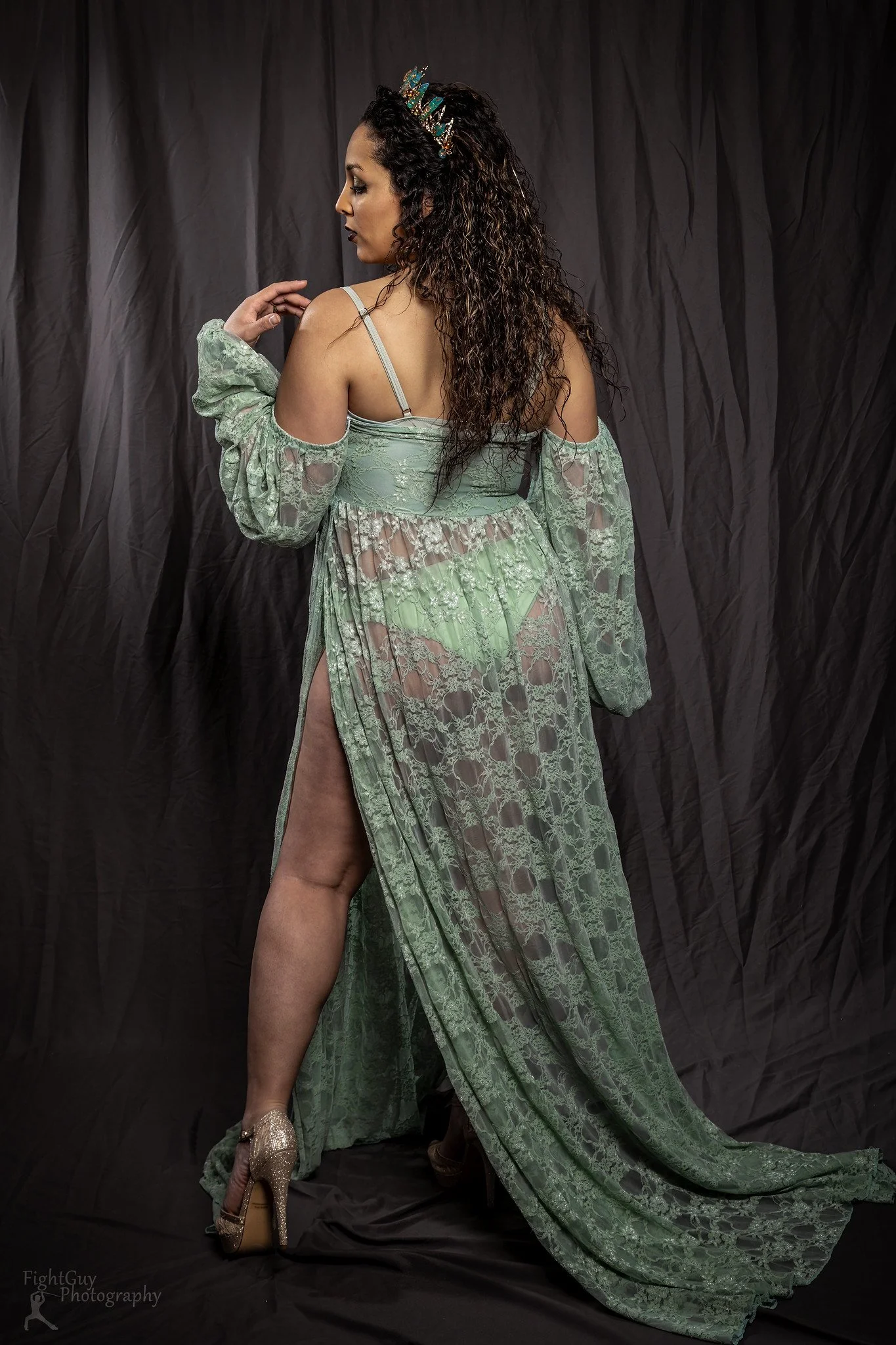 Woman in green lace dress with high heels, standing against black backdrop, looking to the side.