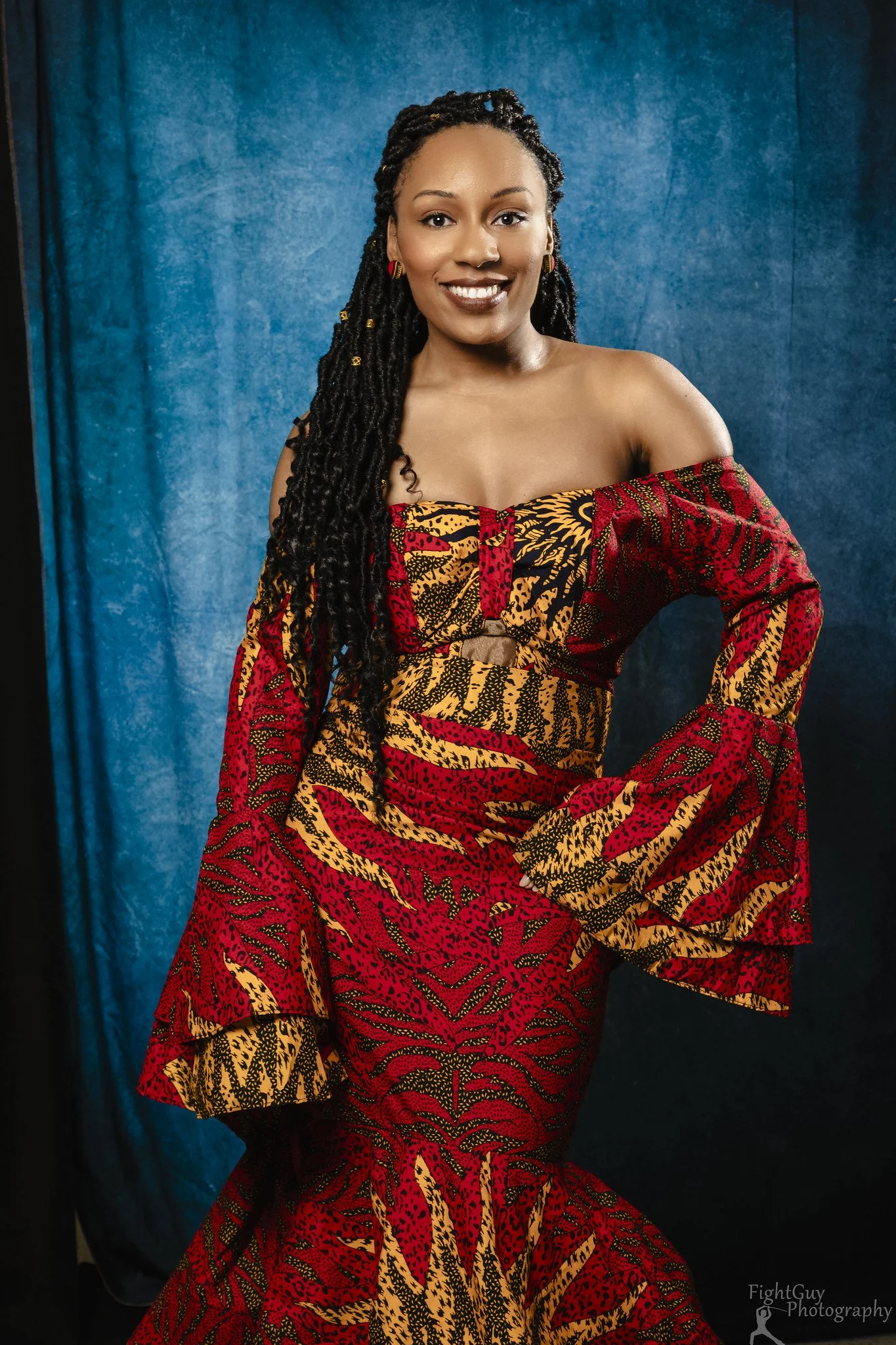 A woman with long dreadlocks wearing a colorful off-shoulder African print dress standing in front of a blue backdrop.