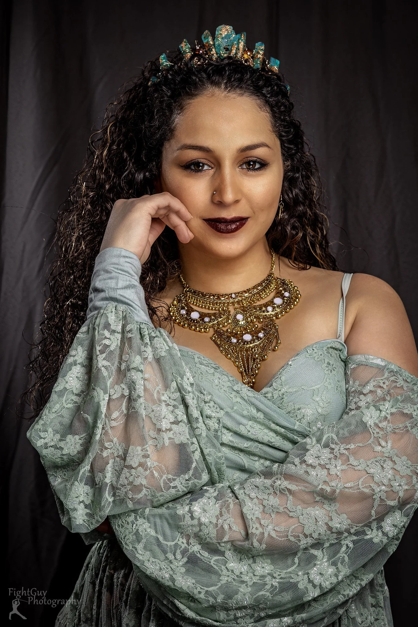 A woman with curly hair wearing a green lace dress, bold dark lipstick, and layered gold jewelry, posing indoors against a dark background.