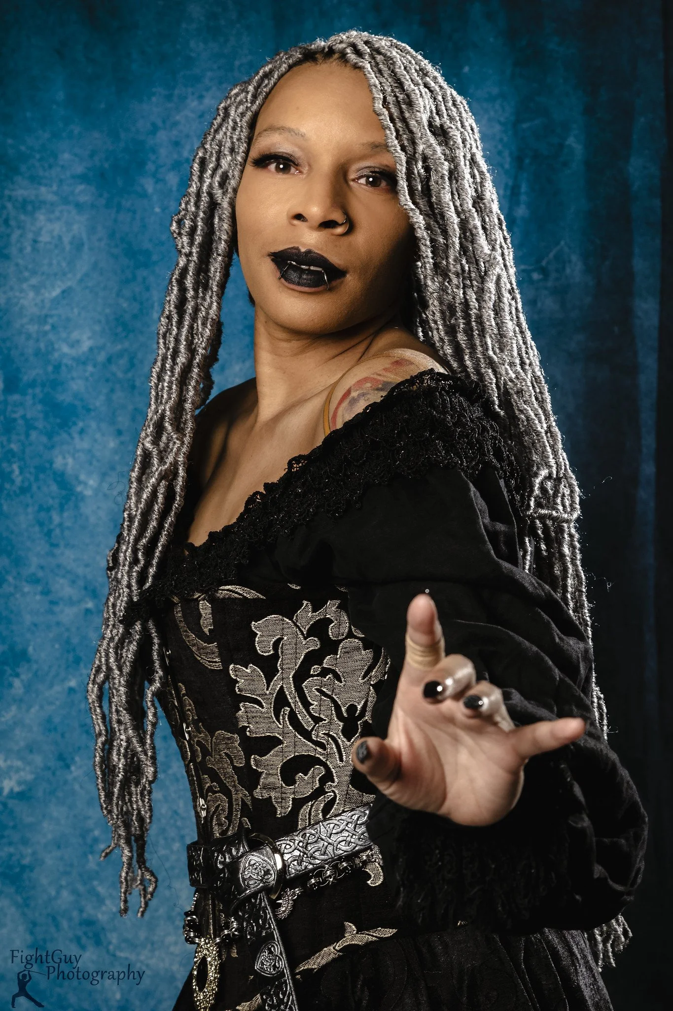 A woman with long gray dreadlocks, black lipstick, and a nose ring poses against a blue textured background, wearing a black and silver ornate gothic-style outfit, extending her hand with fingers slightly curled.