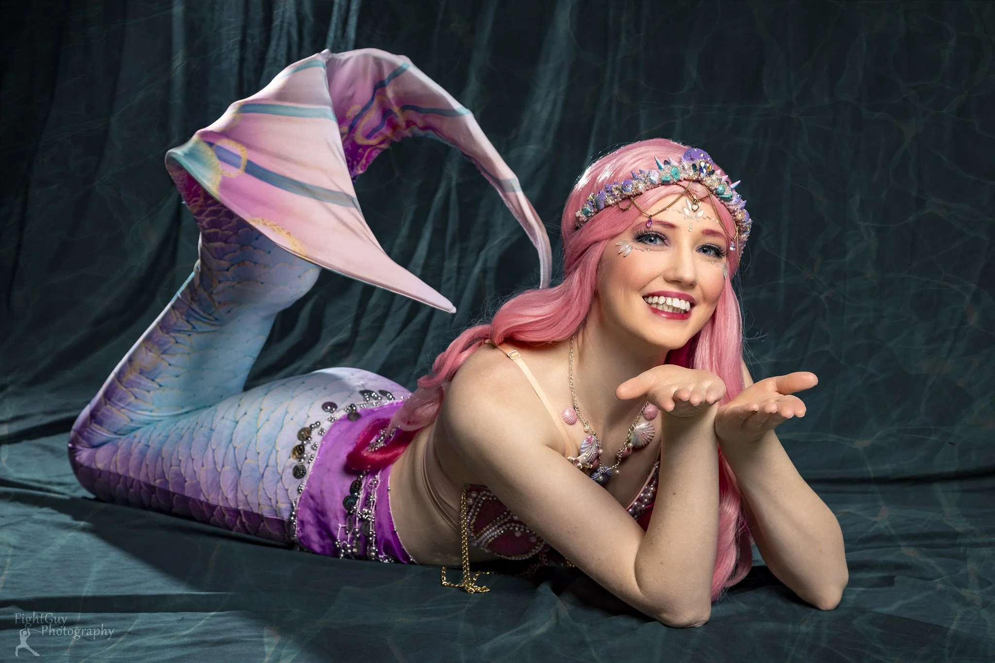 A woman dressed as a mermaid with pink hair, a jeweled crown, and makeup, lying on a dark green fabric backdrop, smiling and blowing a kiss.