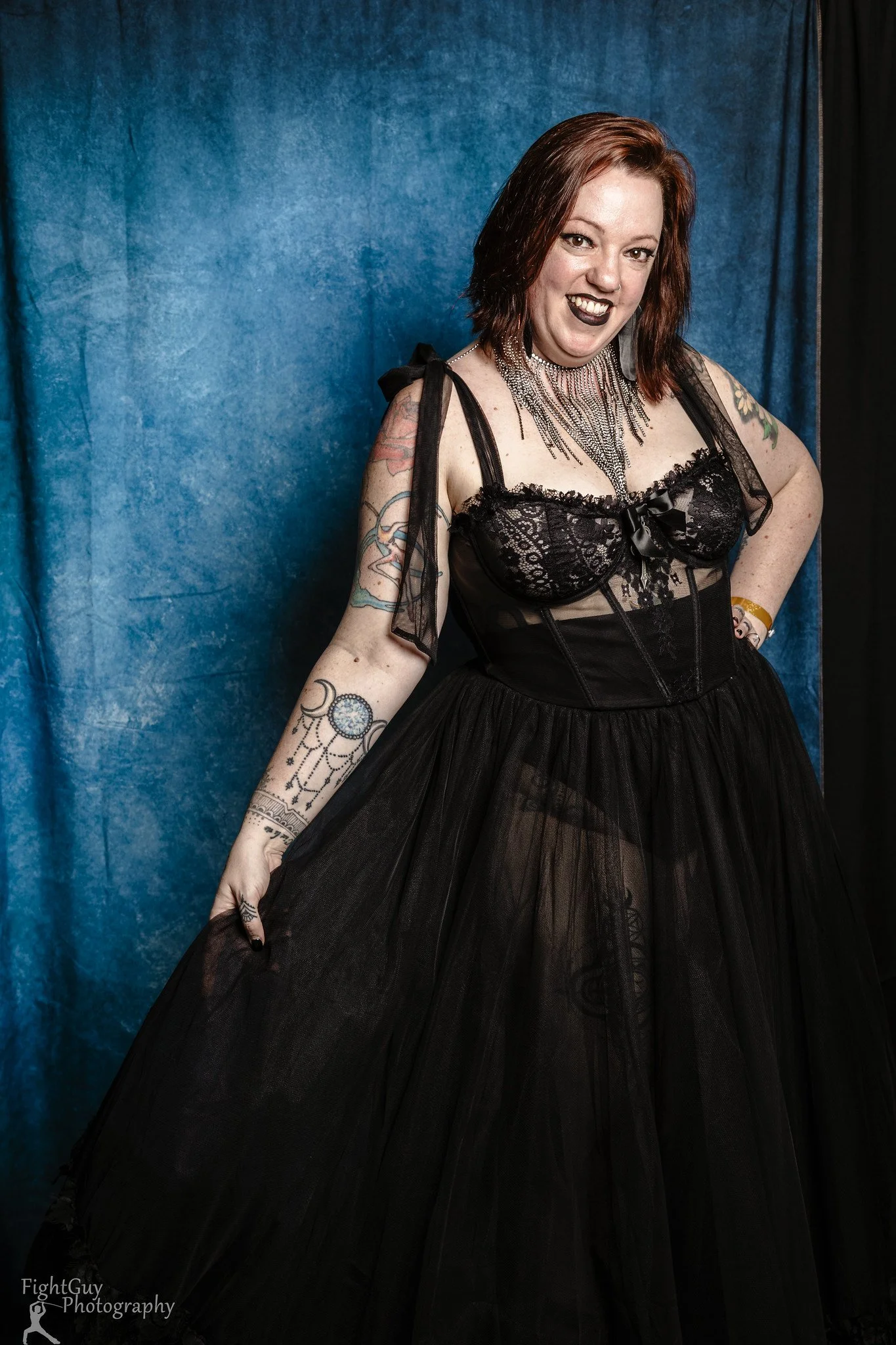 A woman with red hair and tattoos wearing a black lace and tulle dress, smiling in front of a blue textured backdrop.