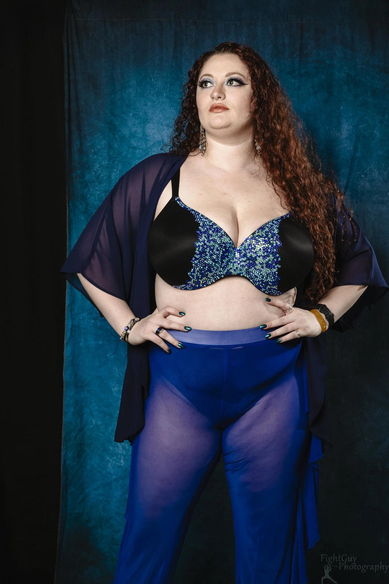 A woman with long curly red hair stands against a dark background, wearing a black and blue bejeweled bra, sheer blue pants, and a matching blue cover-up, with her hands on her hips and a thoughtful expression.