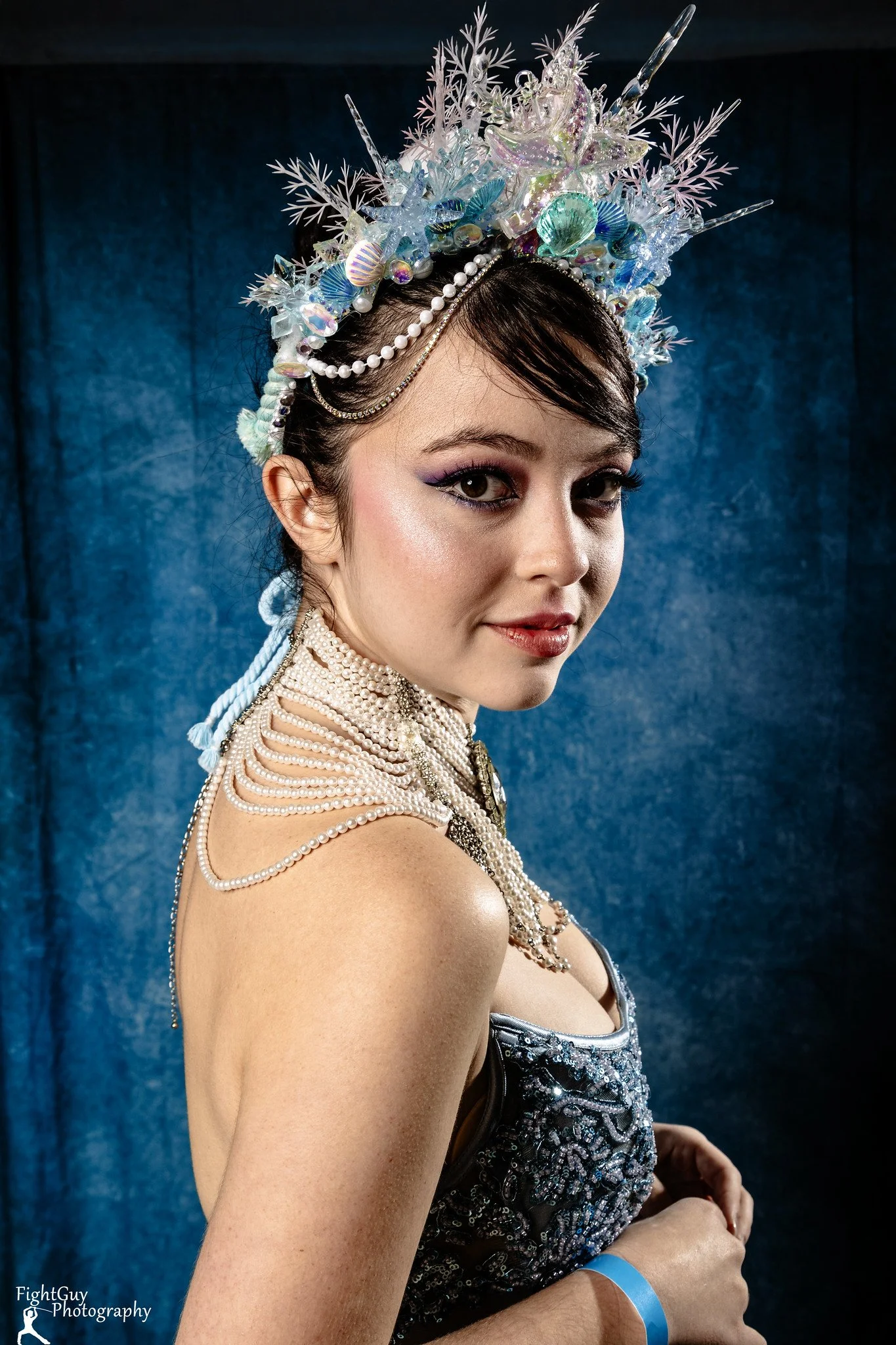 A woman in an elaborate ocean themed headpiece with pearls, seashells, starfish, and coral, wearing a beaded dress and pearl necklaces, standing in front of a blue backdrop.