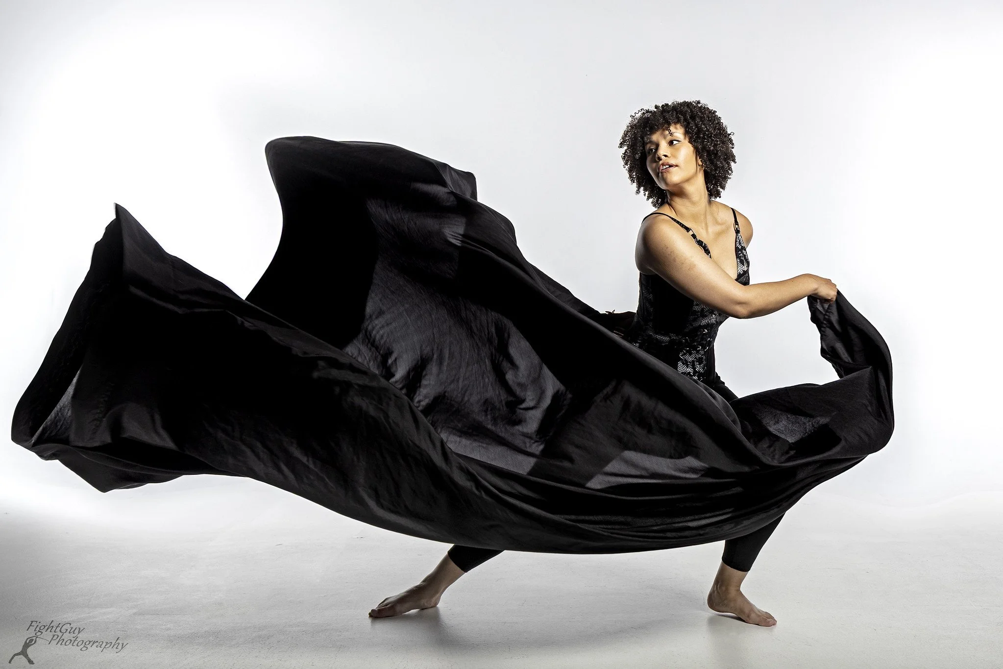 A dancer with curly black hair on a blank white background, she's moving a black silk sheet across the frame, it billows out in front of her.