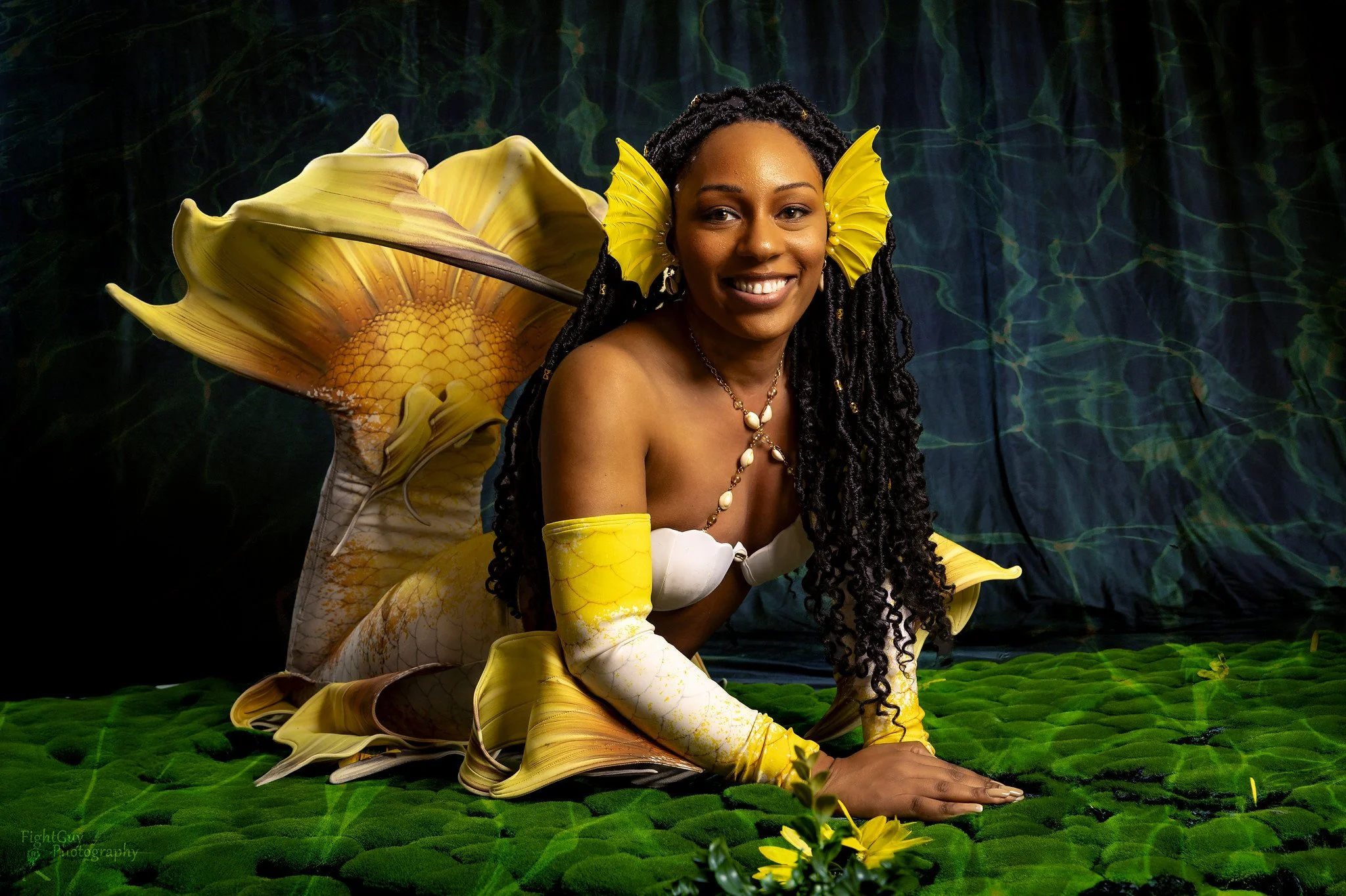 A woman dressed in a yellow and white floral-inspired costume with large yellow flower-shaped earrings and jewelry, posing on a green textured surface with a dark backdrop featuring a large flower design.