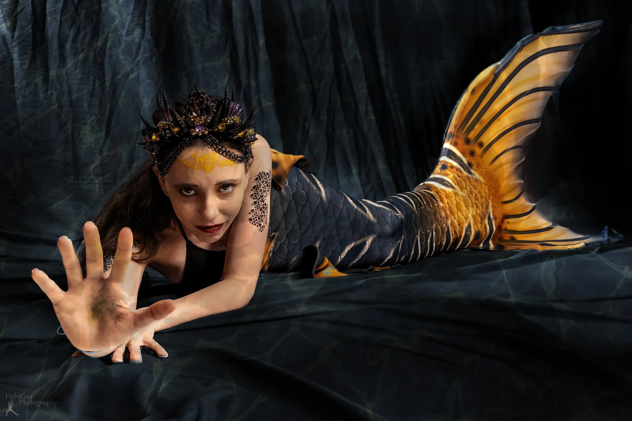 Woman dressed as a mermaid with a fish tail, reaching forward with her hand, posed on dark fabric with a forest background.