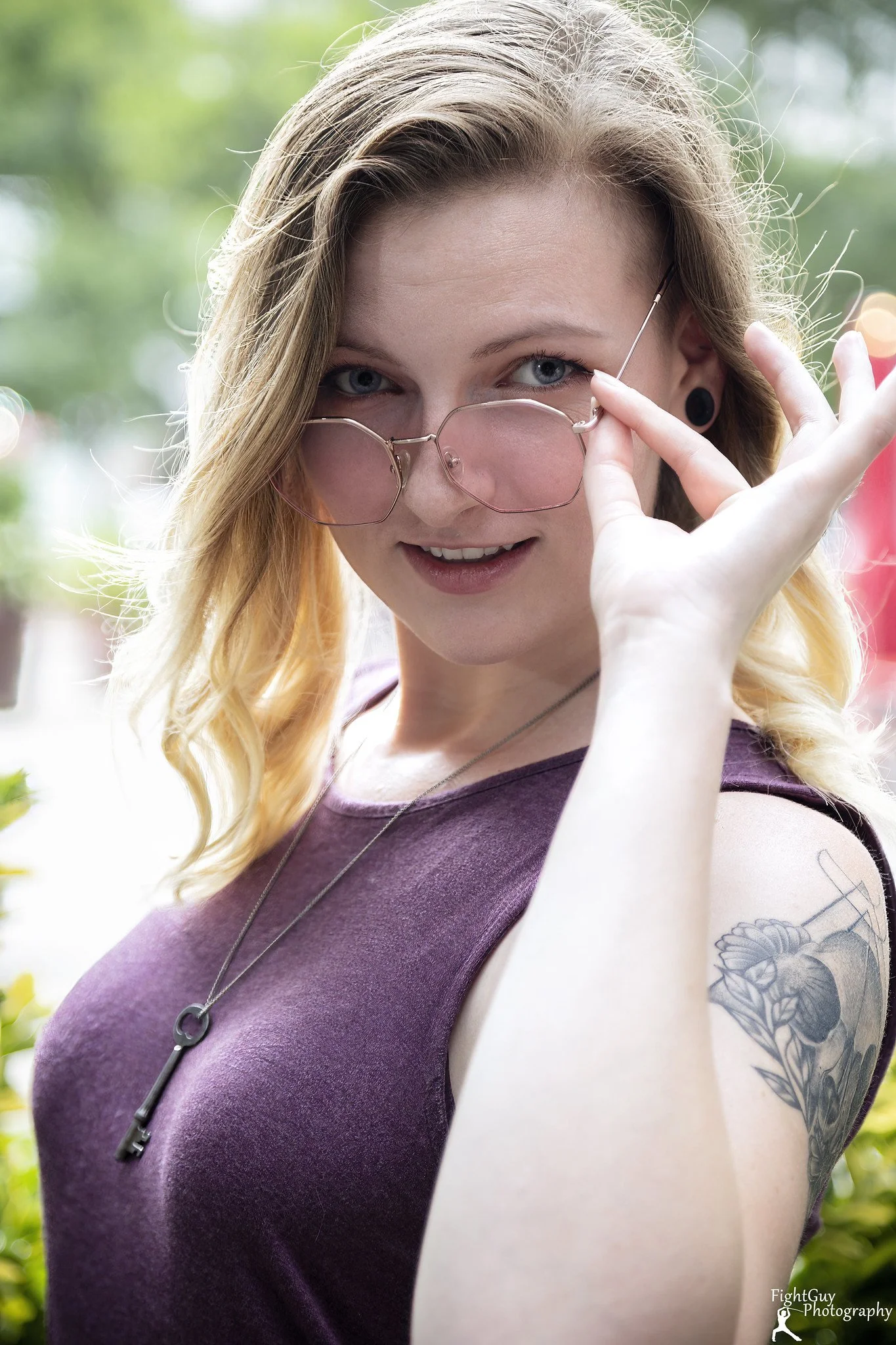 A young woman with blonde hair, wearing pink-tinted glasses, a purple sleeveless top, and a necklace with a key pendant, is outdoors with greenery in the background, smiling and looking at the camera.