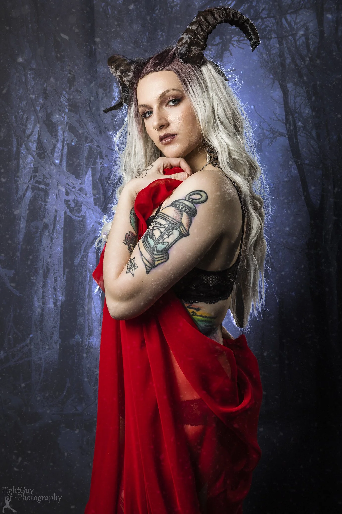 A woman with horned headpiece, tattoos, and long blonde hair stands against a dark, misty forest background, holding red fabric.