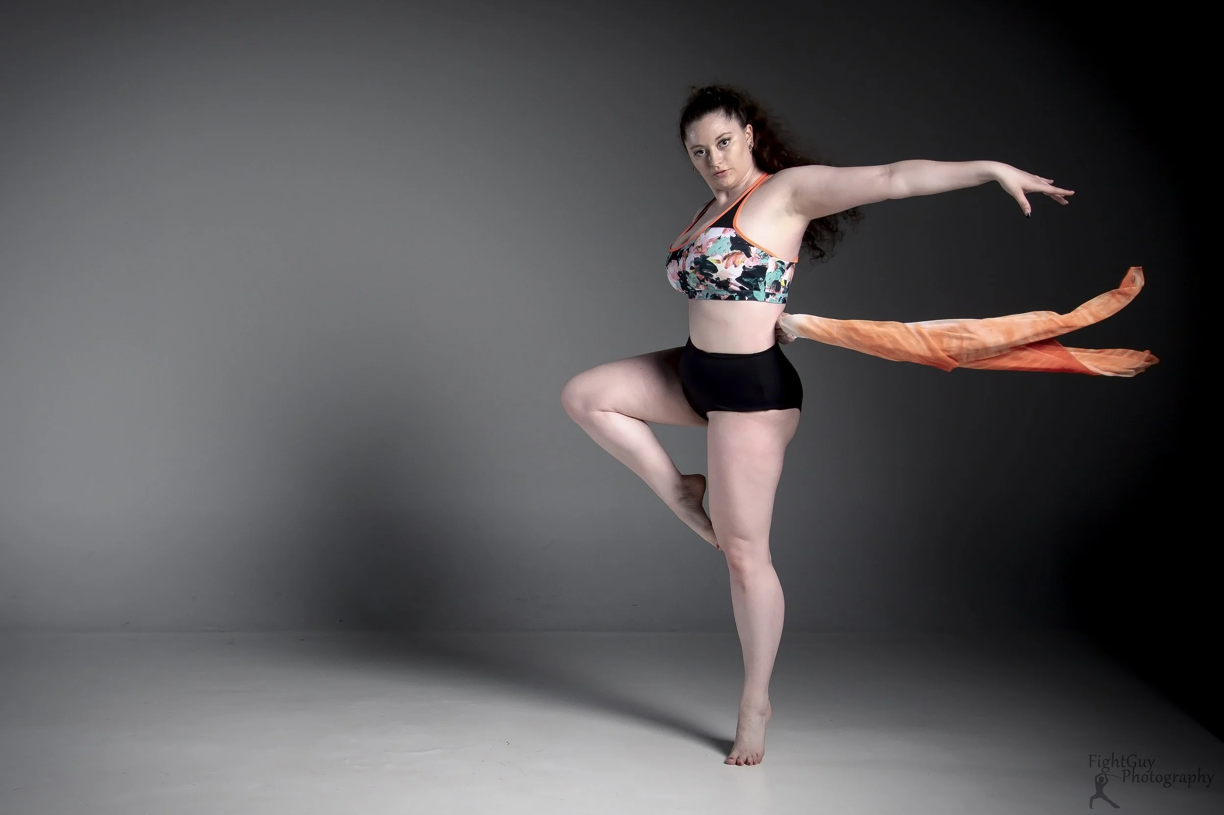 A woman in athletic clothing poses with one leg raised and arms extended to the sides against a gray background.