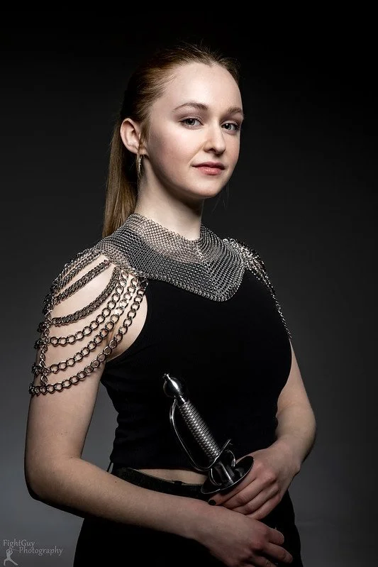A young woman with blonde hair tied back, wearing a black outfit with silver chain embellishments on her shoulders and neckline, holding a sword with a black handle, against a dark background.