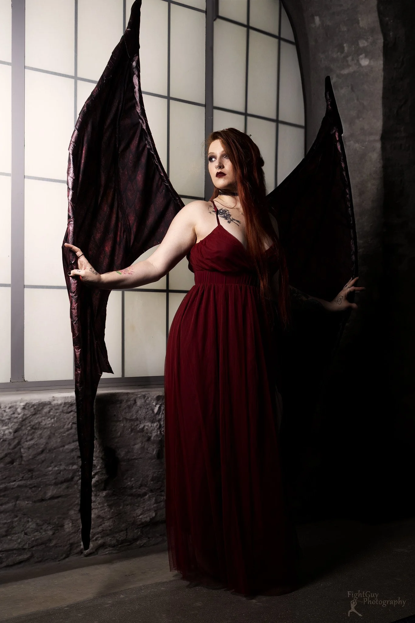 A woman with long red hair, tattoos, and dark makeup wearing a flowing red dress and black bat wings posing in front of a window with grid panes.