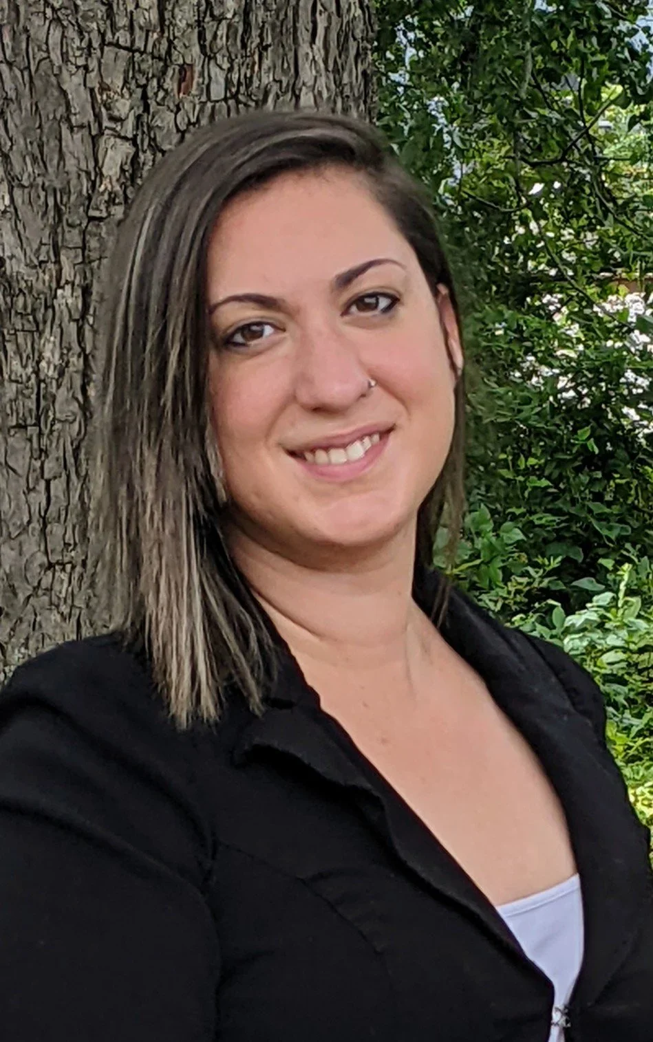 A woman with shoulder-length brown hair, wearing a black jacket over a white top, standing outdoors next to a large tree and green shrubs, smiling at the camera.