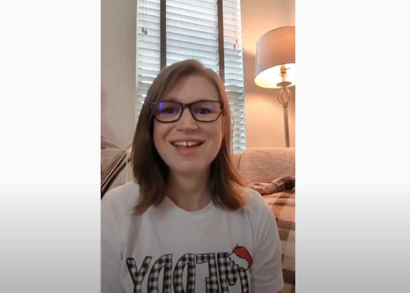 A person with glasses and brown hair is smiling while sitting indoors. They are wearing a white shirt with text on it. Behind them is a window with blinds, a floor lamp, and part of a couch covered with a plaid blanket.