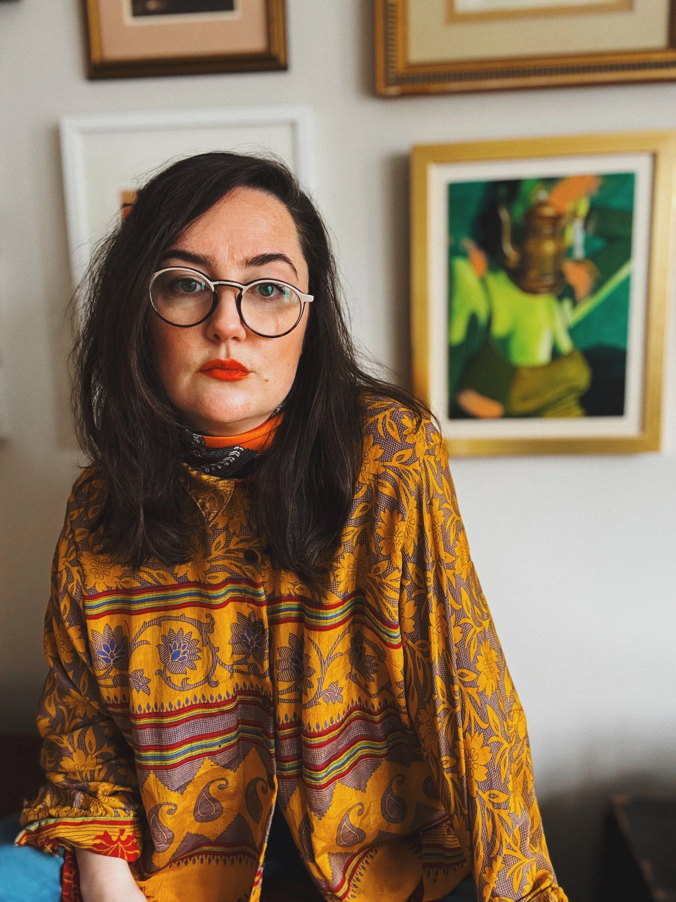 A woman with dark hair and glasses is wearing a patterned, colorful shirt and orange lipstick, standing in front of a wall decorated with framed artwork.