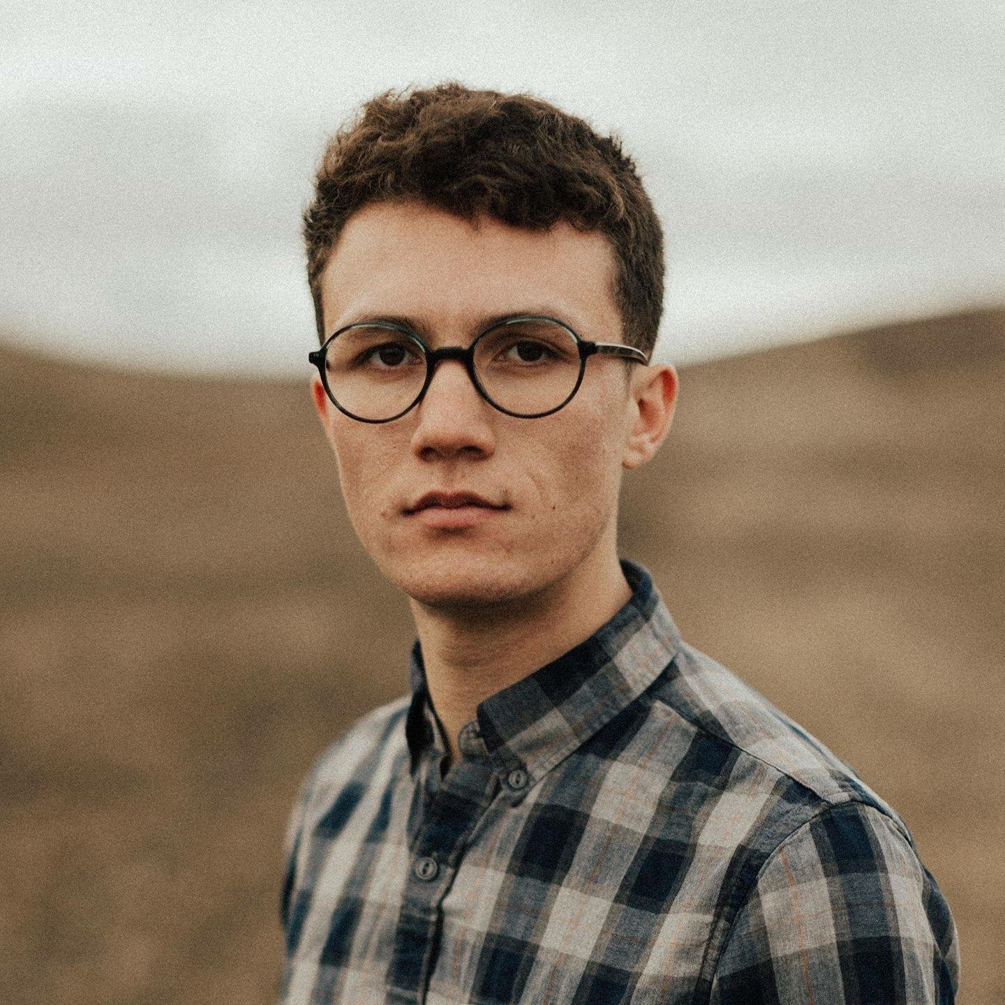 Portrait of a young man with short curly hair and round glasses, wearing a plaid shirt, standing outdoors with a blurred landscape in the background.