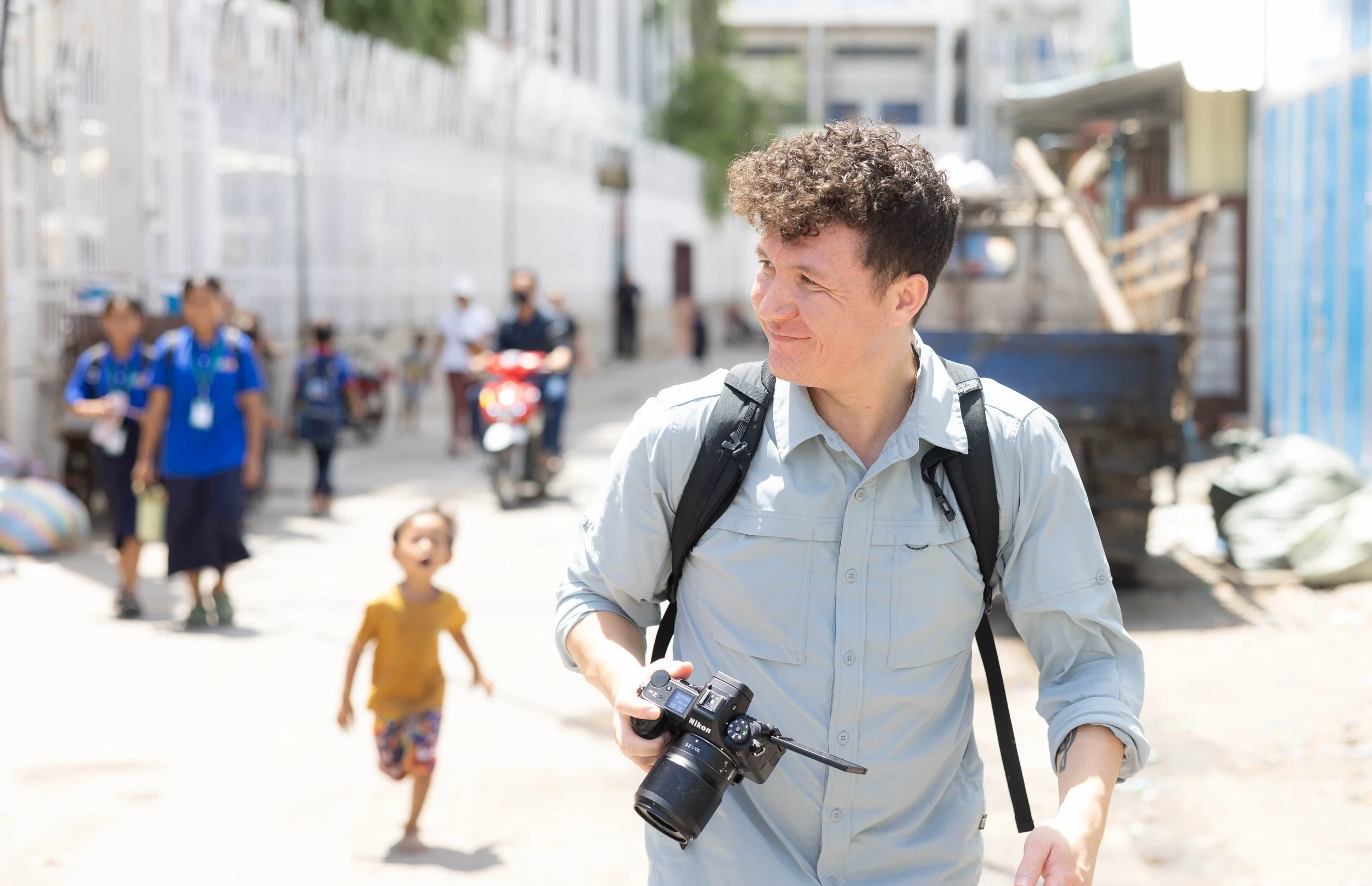 Man holding a camera smiles as he walks down a busy street with children chasing him and a man on a motorcycle.