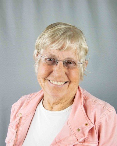 Smiling elderly woman with short blonde hair, wearing glasses, a white shirt, and a light pink jacket.