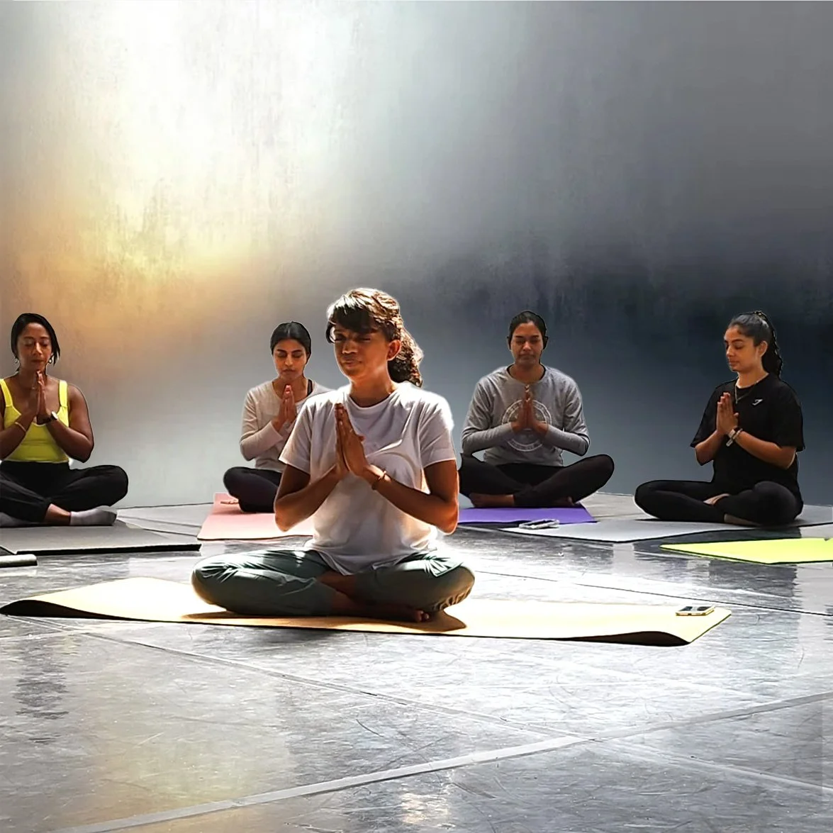 Group of five women practicing yoga or meditation, seated on mats in a cross-legged position with hands in prayer pose, in a dimly lit indoor studio.