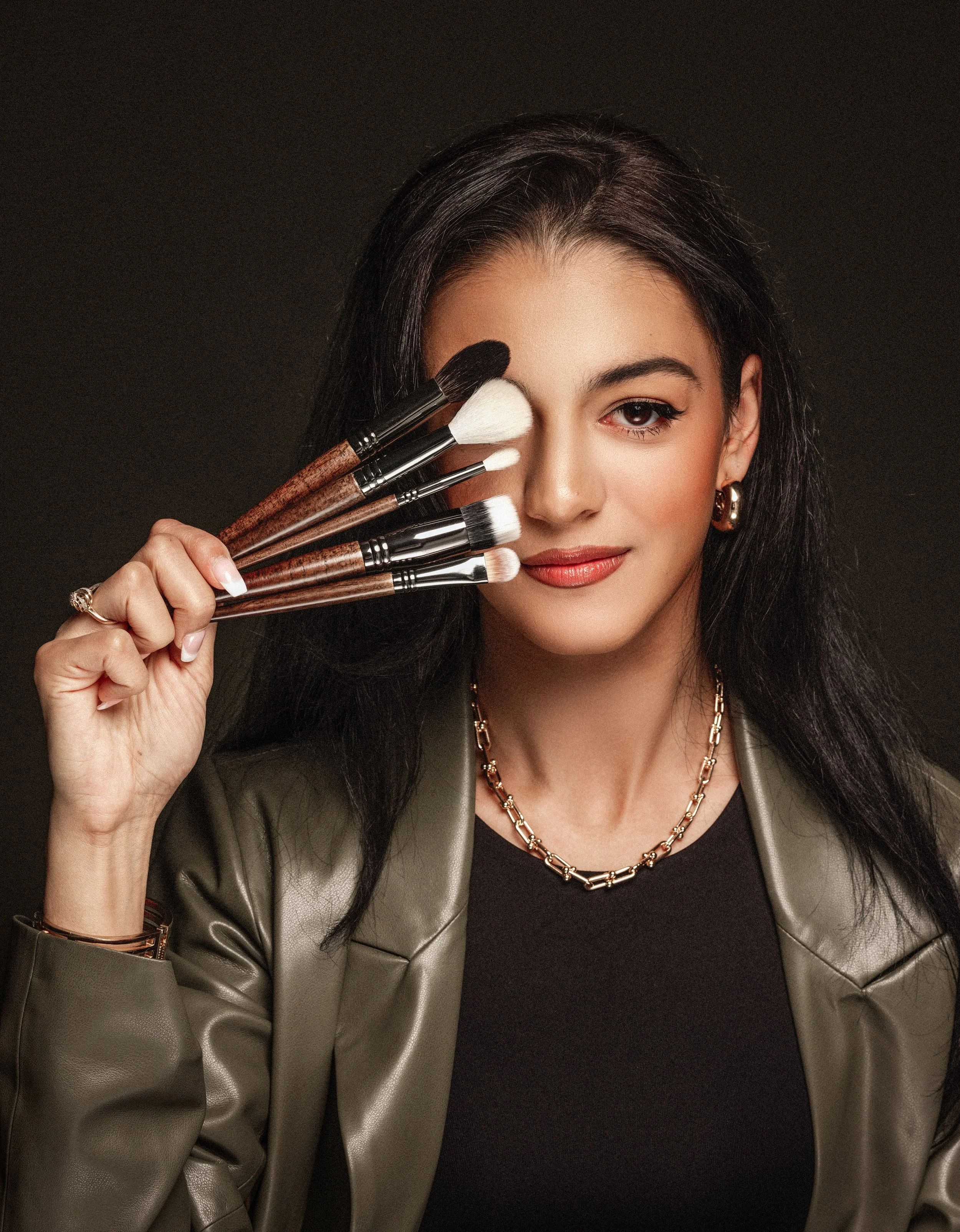 A woman with dark hair and jewelry holding makeup brushes in front of her eye.