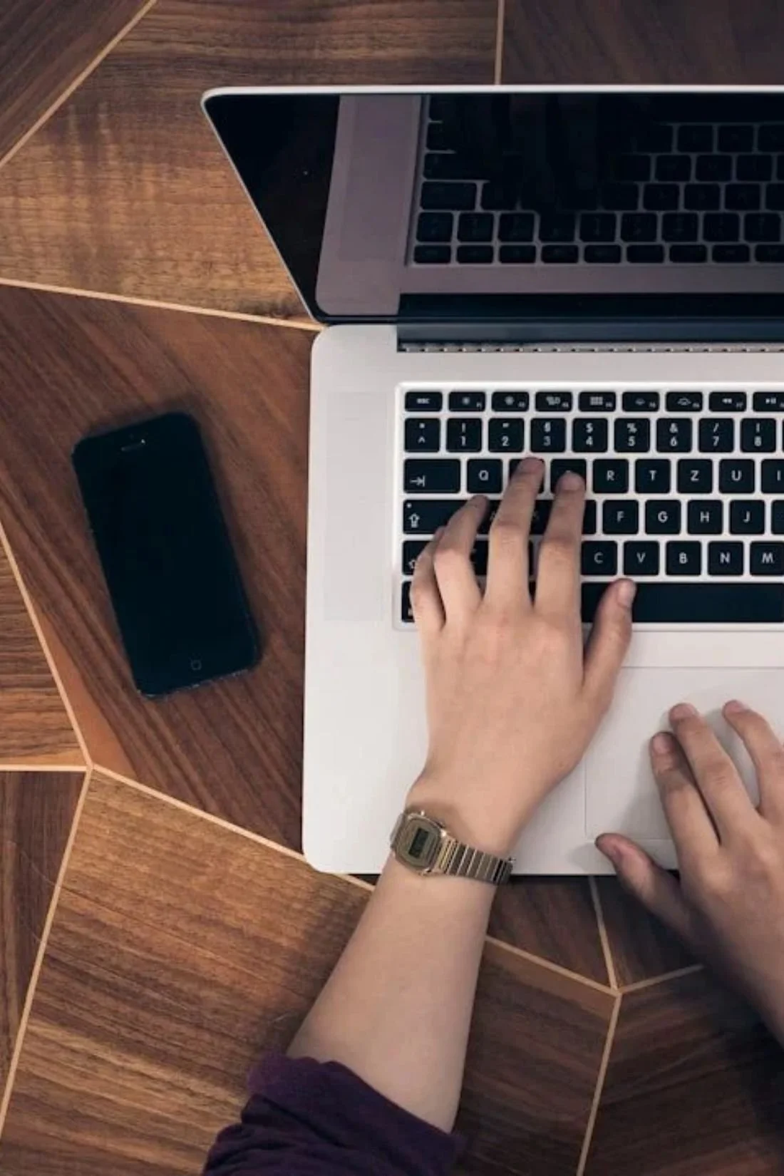 Top-down view of a person typing on a silver laptop with a black keyboard, on a wooden table.