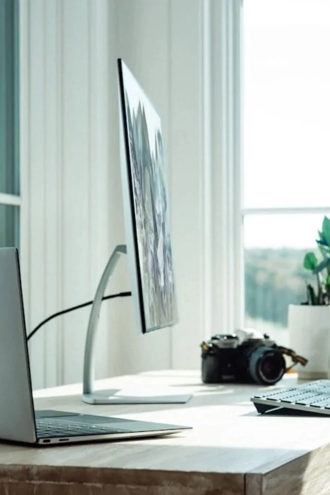 An organized home office desk by a large window with a computer monitor, a laptop, a camera, a keyboard, a mouse, potted plants, and mugs.