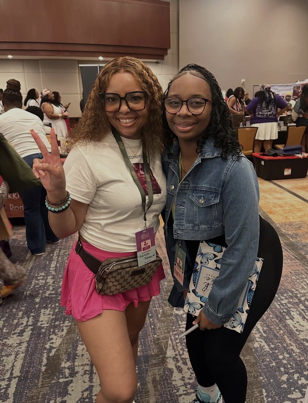 Two young women with glasses smiling at a conference or event. One shows a peace sign with her fingers. They are surrounded by other attendees and event booths.
