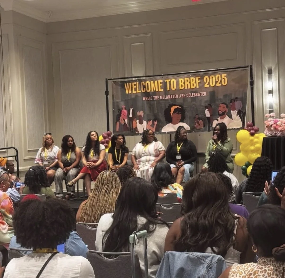 A panel of seven women seated on stage at the BRBF 2025 event, with a large banner behind them reading "Welcome to BRBF 2025" and "Where the Melanated are Celebrated". Attendees are seated in front of the stage in a conference room decorated with yel