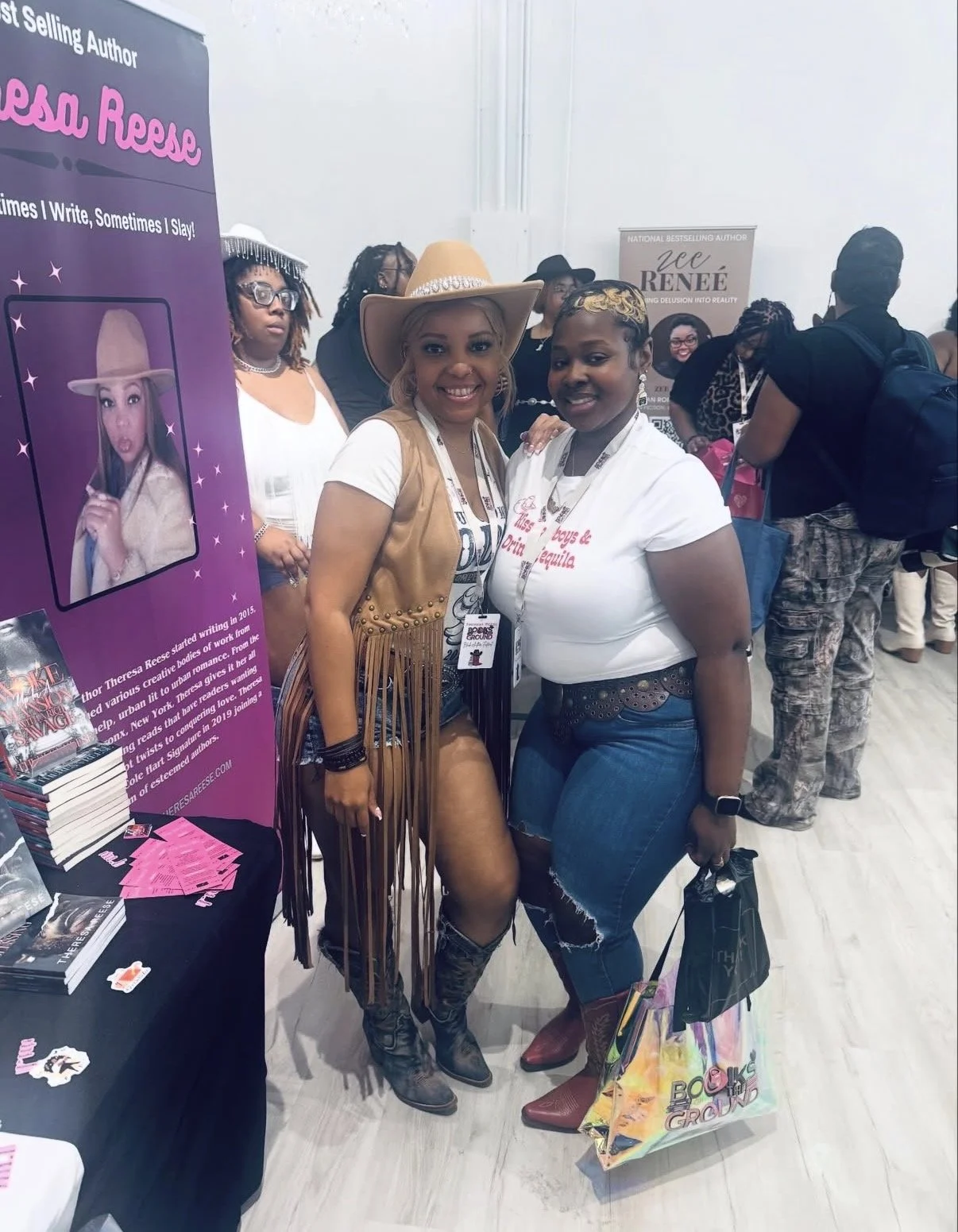 Two women posing together at a book event, standing in front of a purple promotional display for author Theresa Reese. The woman on the left wears a tan cowboy hat, tan vest, and cowboy boots, while the woman on the right wears a white T-shirt and je