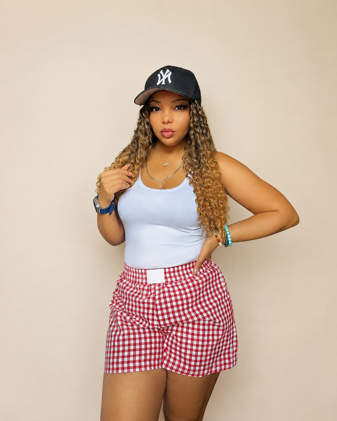 A young woman with long, curly blonde hair, wearing a black New York Yankees baseball cap, white tank top, red and white checkered shorts, and colorful jewelry, standing against a plain beige wall.