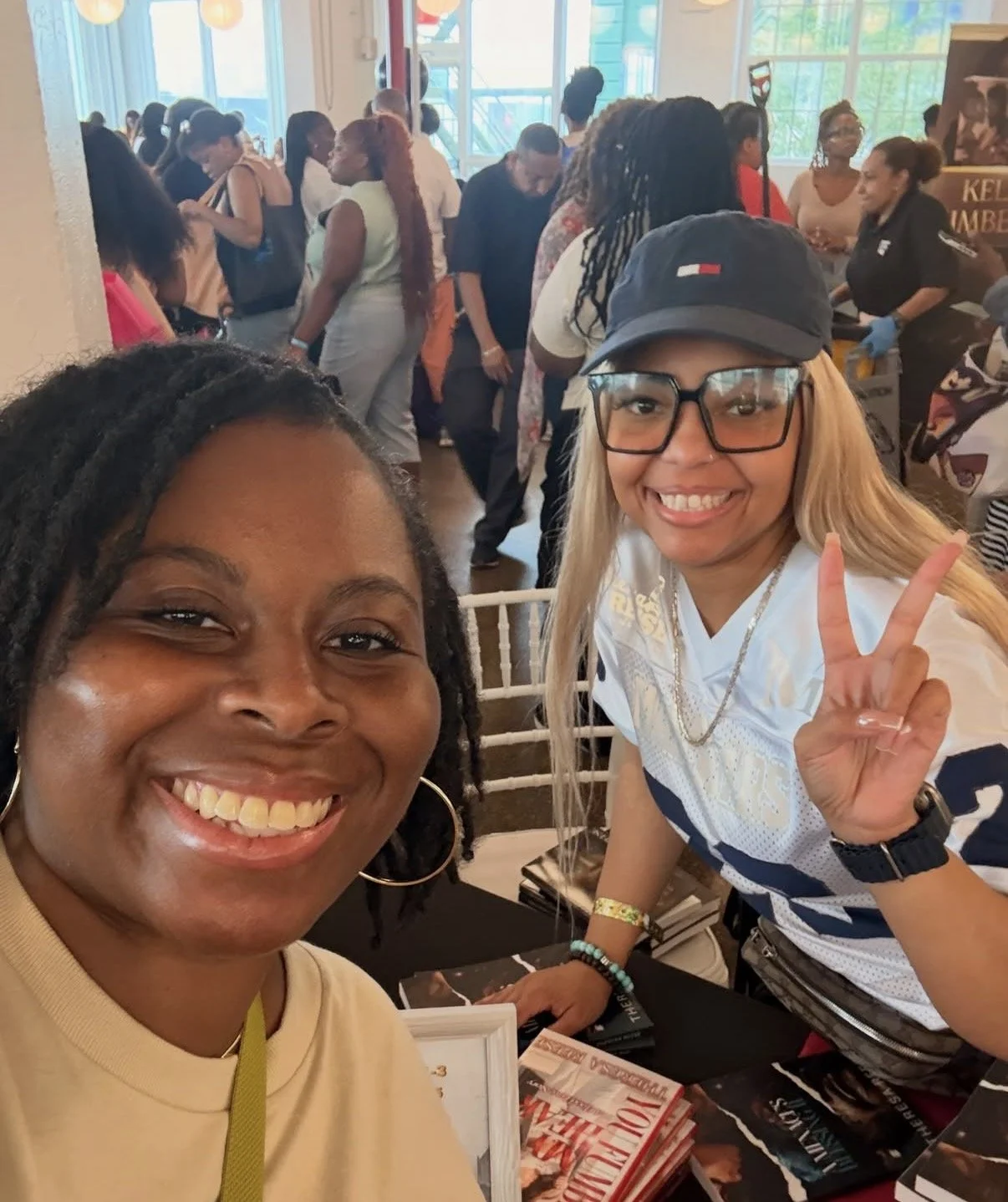 Two women smiling for a selfie at a crowded table during a book event or signing, with one making a peace sign. In the background, people are mingling and browsing books inside a well-lit room with large windows.