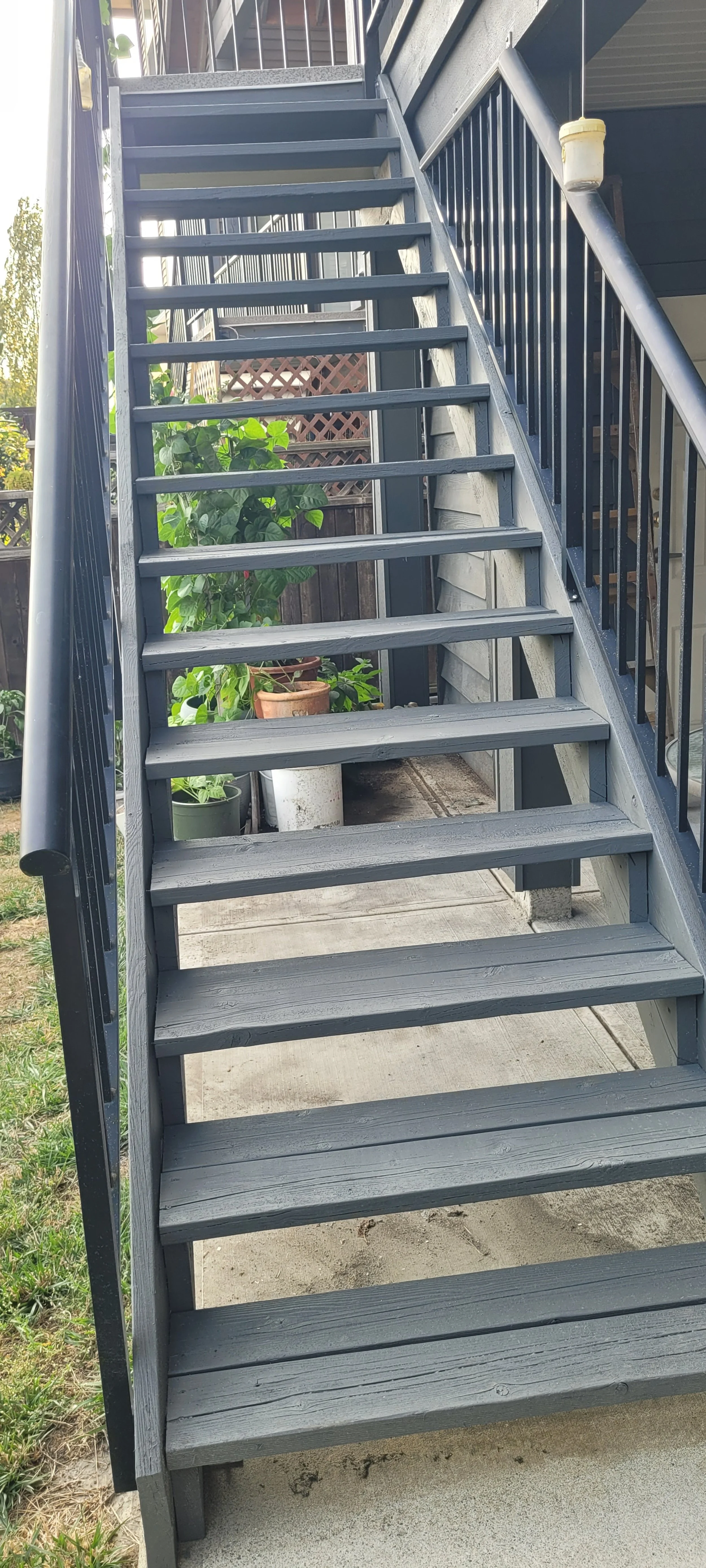 Outdoor gray wooden staircase with black metal railing leading to an upper floor of a building, with potted plants and a wooden fence in the background.