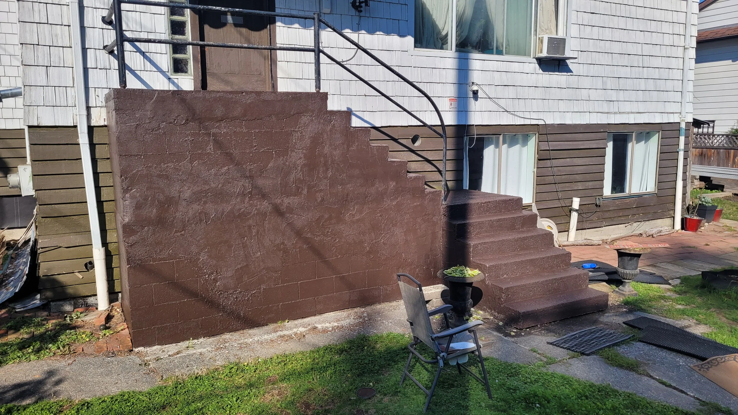 Backyard patio area with steps painted dark brown, a black chair with a white paper, potted plants, and a house with grey and brown siding and propane tank.