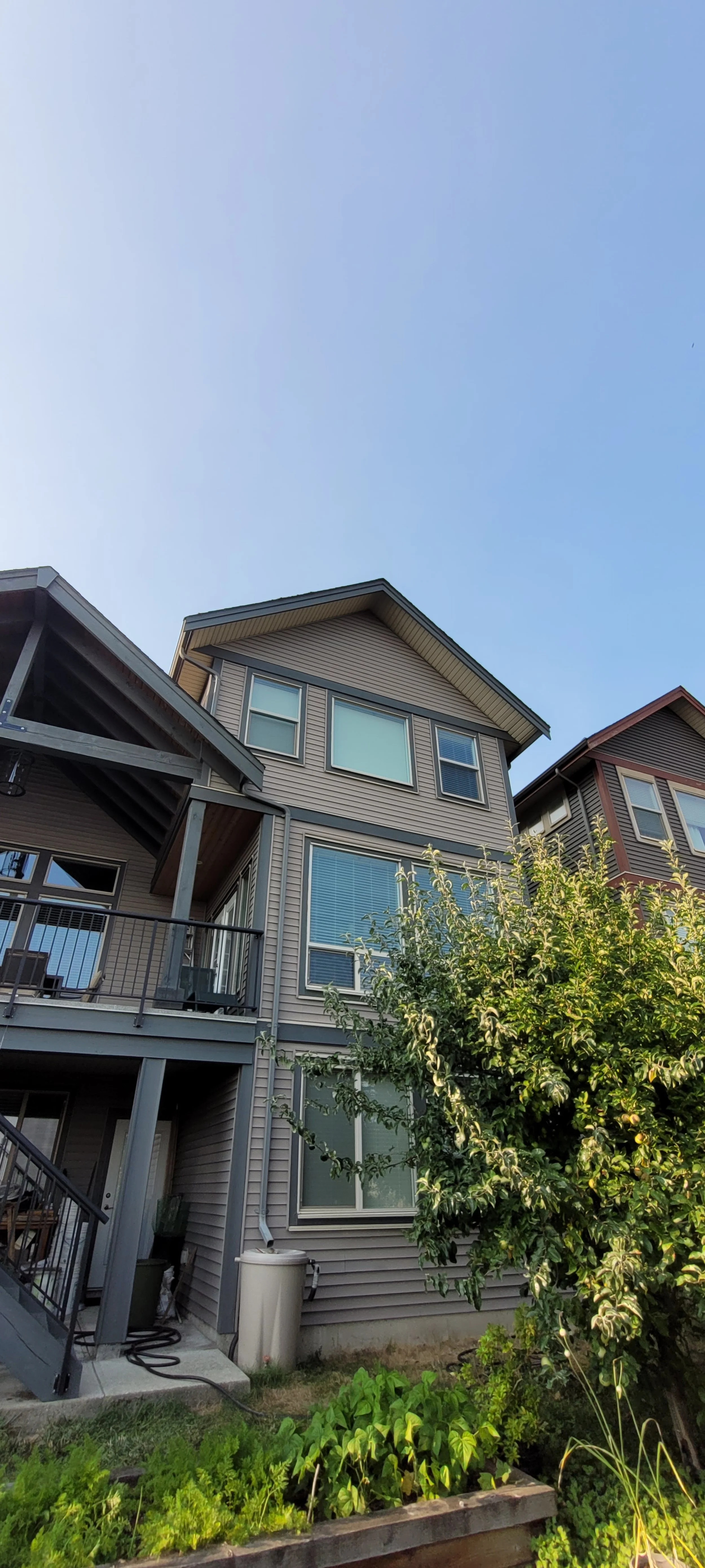 A three-story residential building with beige siding, large windows, a small balcony, and a garden with green plants and a tree in the foreground.