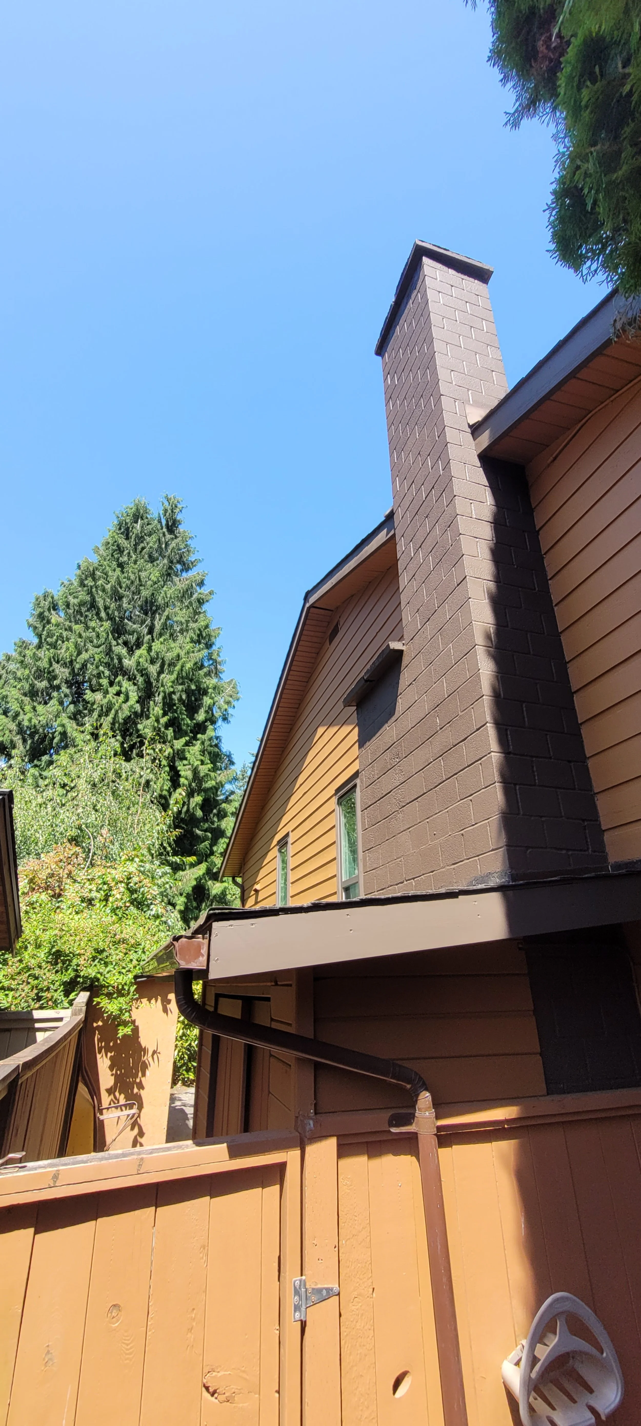 Close-up of a house with a brown brick chimney, tan siding, and a wooden fence, with tall green trees and a clear blue sky in the background.