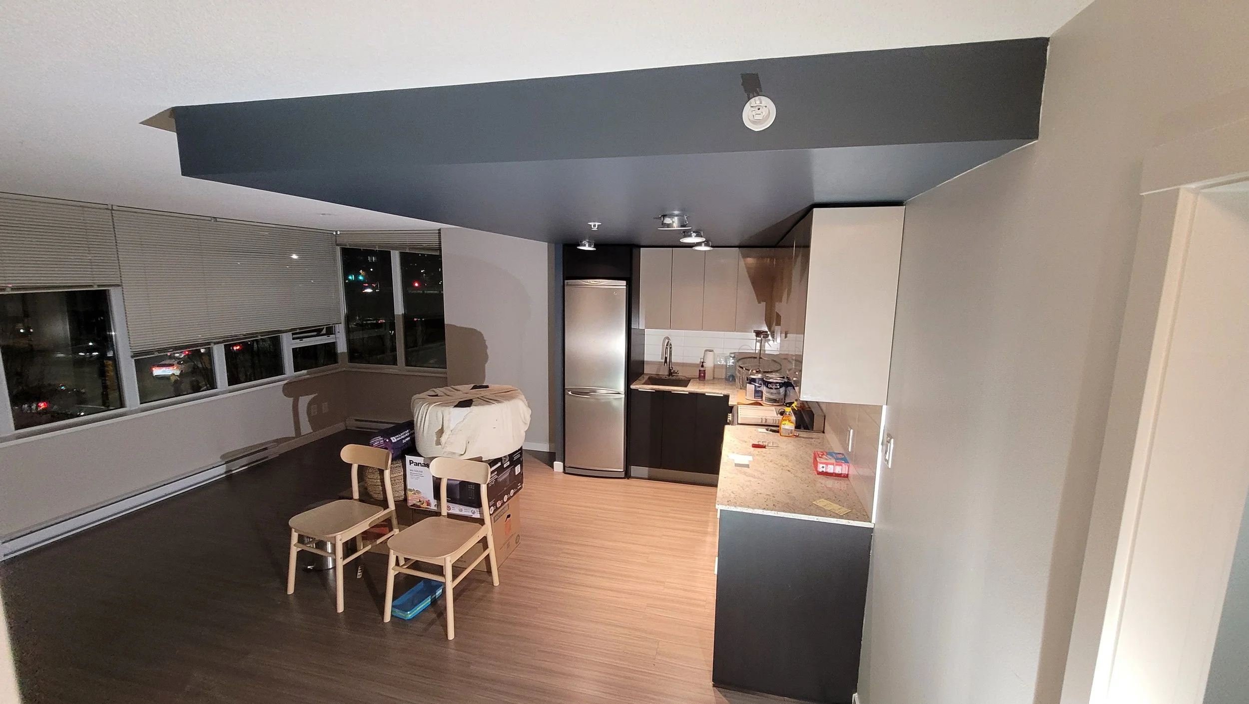 Interior view of an apartment with a kitchen and living area, featuring a refrigerator, chairs, and boxes, with windows showing a night cityscape.