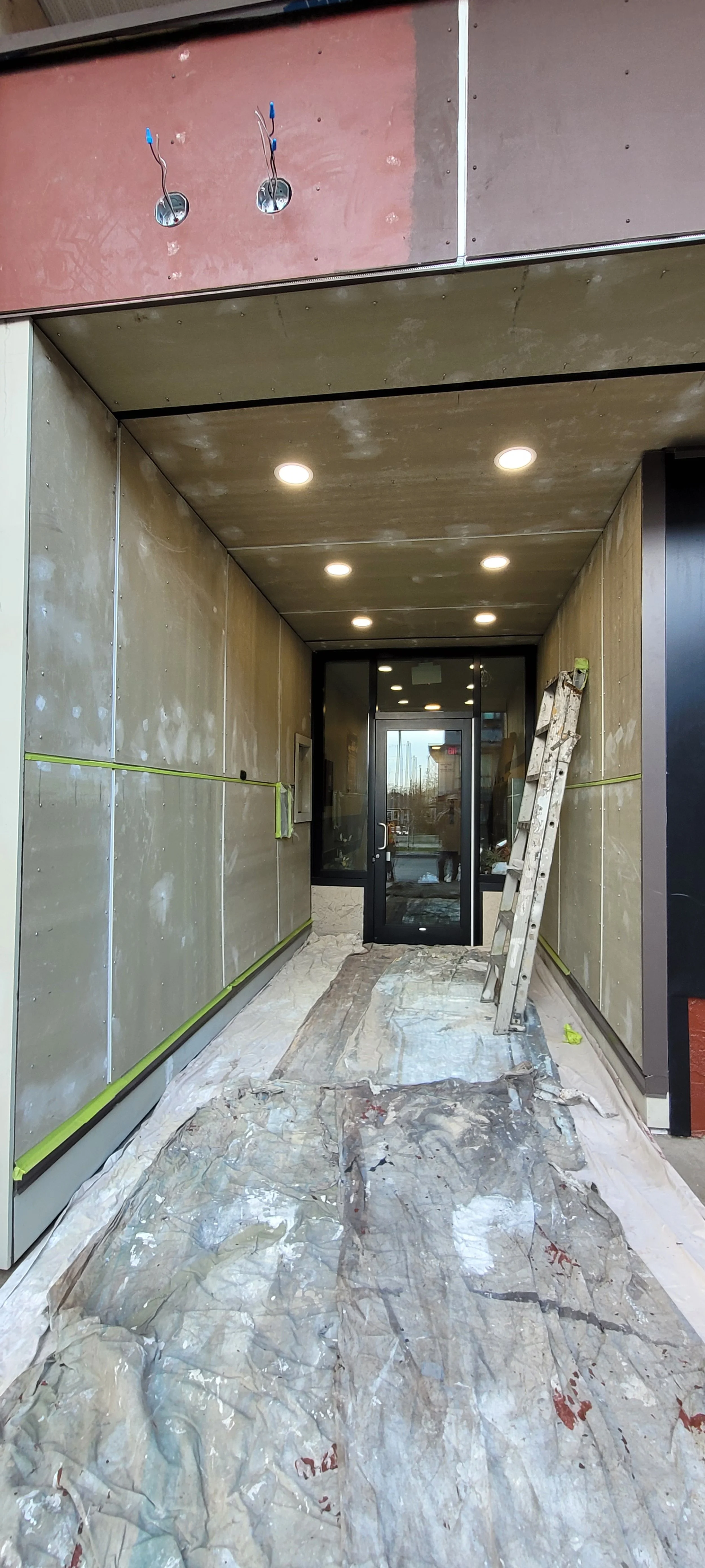Construction site of an entrance with a door at the back, ceiling and walls installed, ladder leaning against the right wall, protected floor covering in place.