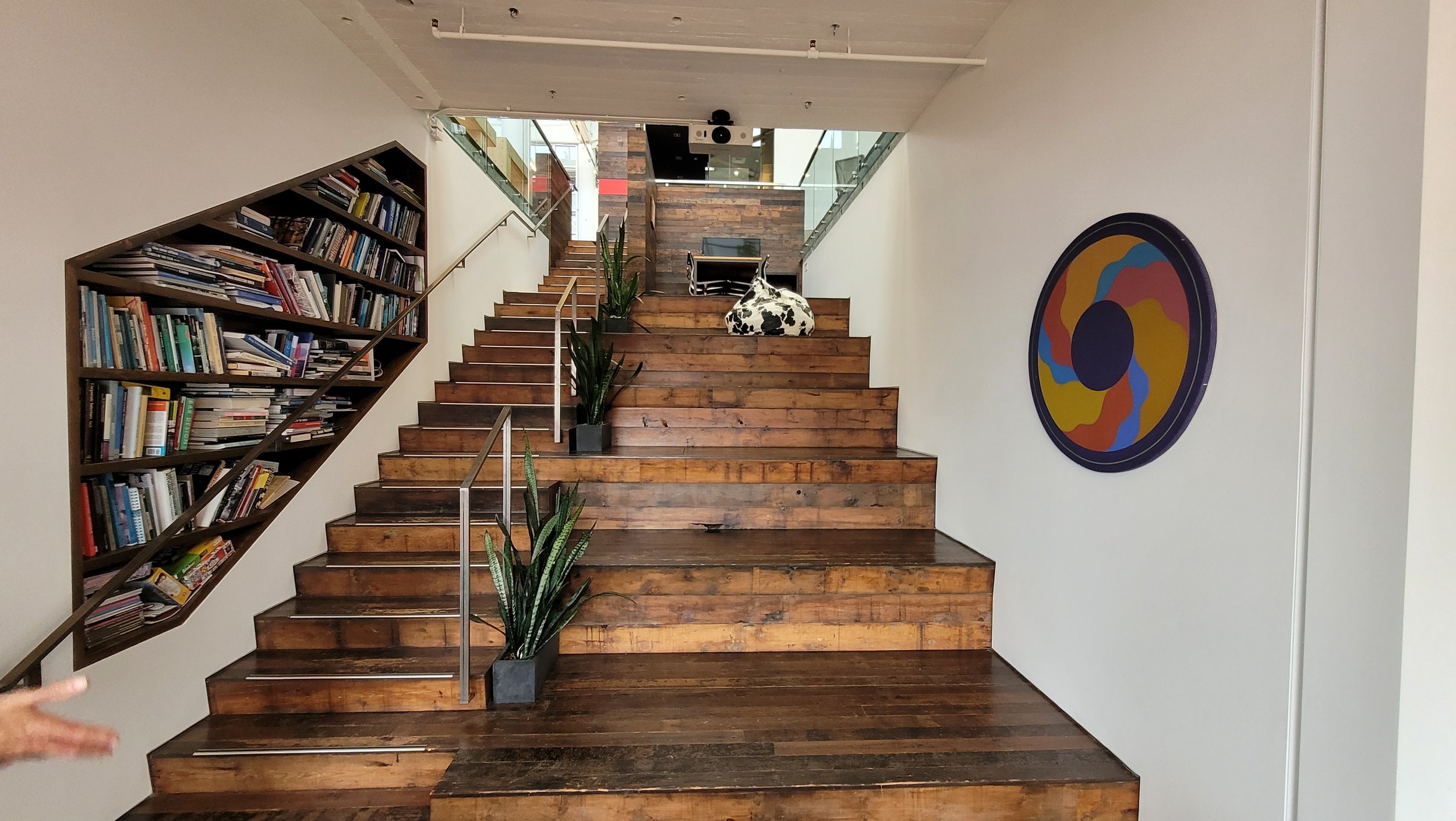 Indoor staircase with wooden steps, black and white cow print cushion on top, potted snake plants on the sides, glass railing, and abstract artwork on the right wall.
