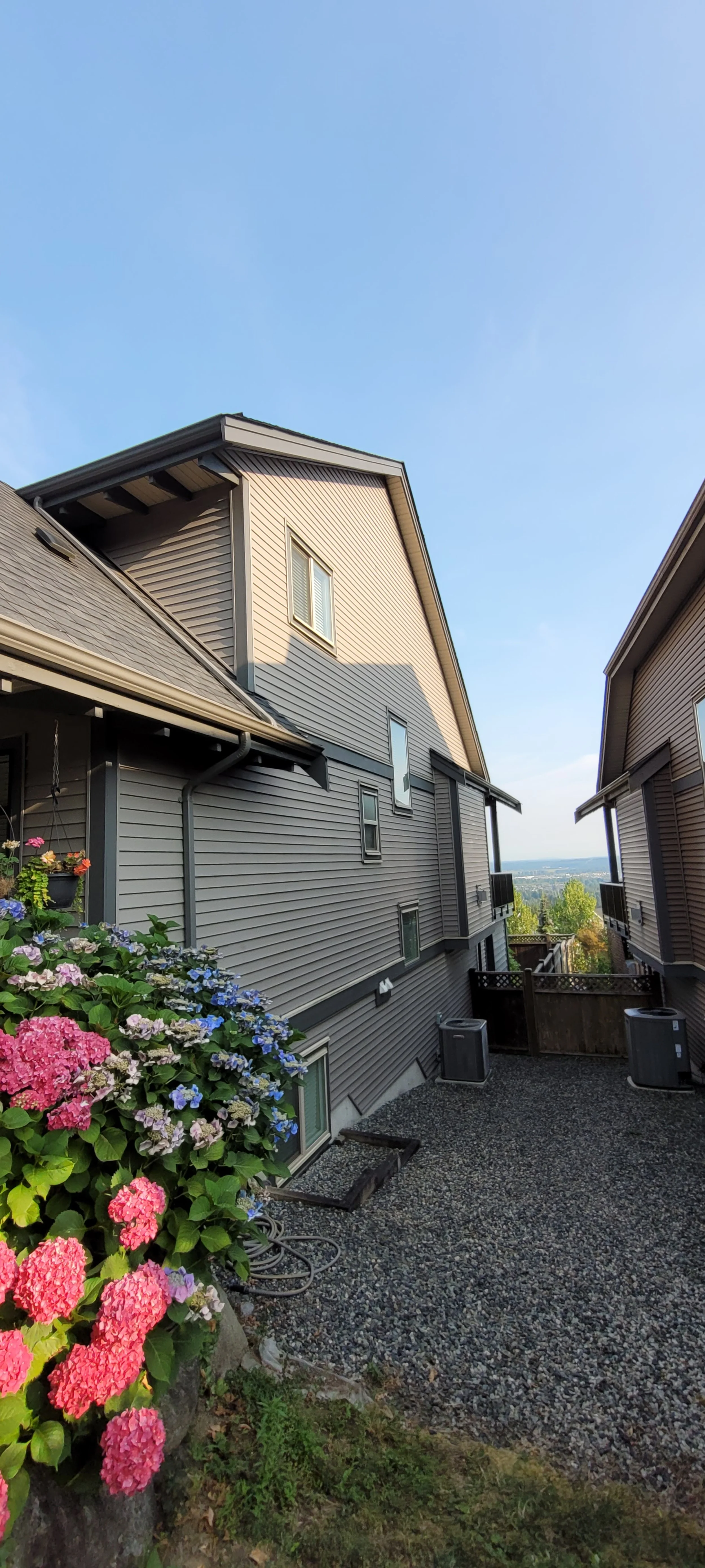 View of two houses with vinyl siding and a small gravel yard, flowers on the left, with a clear sky above.