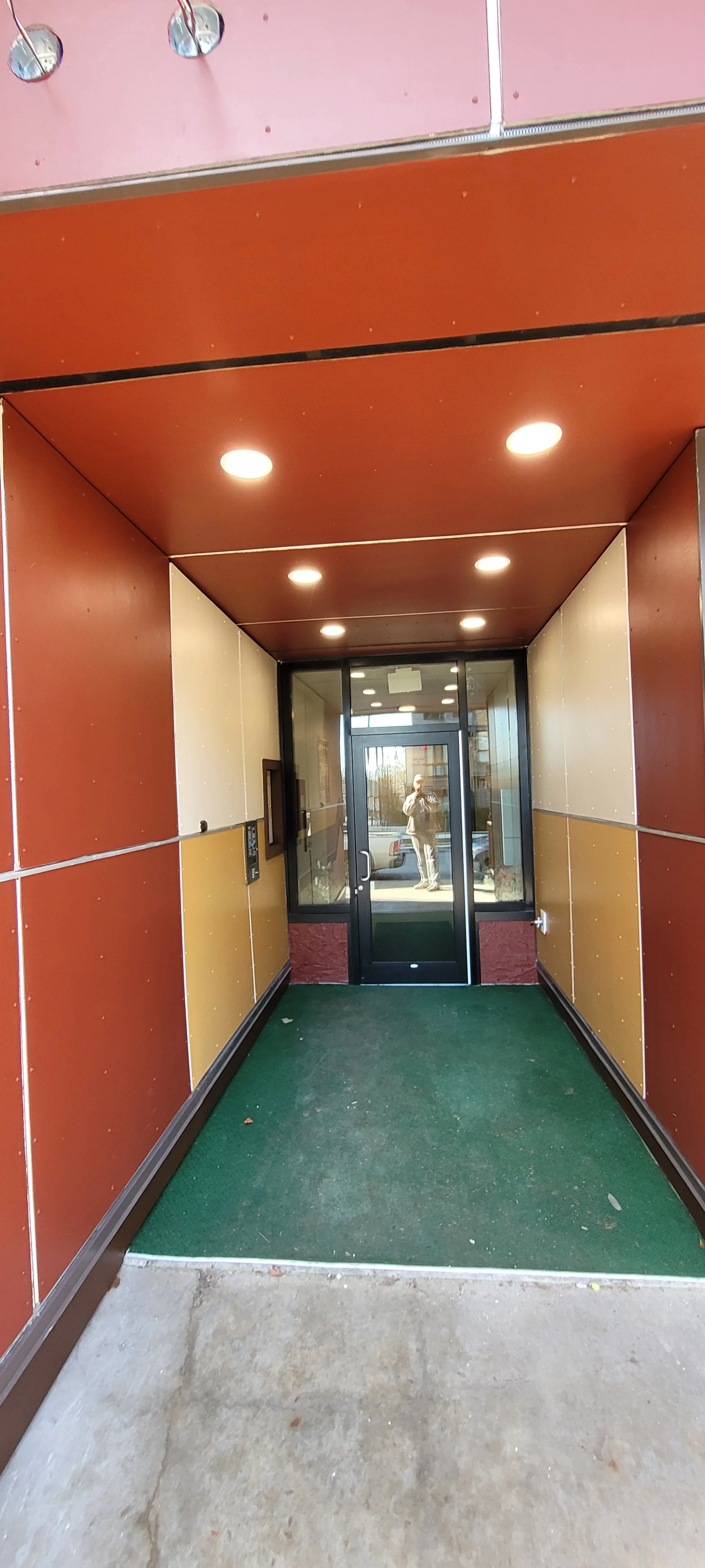 Entrance to a building with a glass door, green carpet, and beige and red walls, reflecting a person taking the photo.