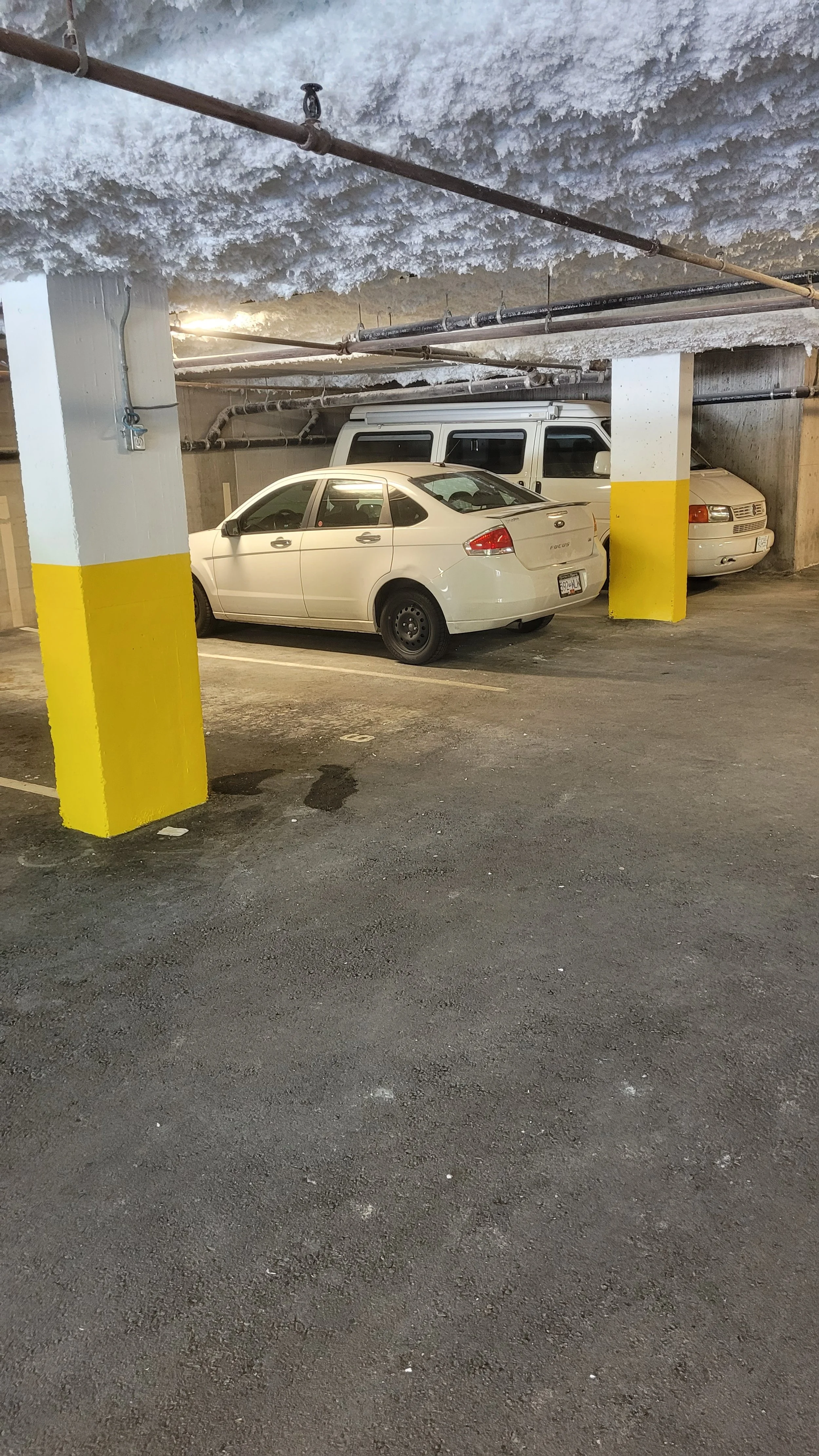 Underground parking garage with two parked vehicles, a white sedan and a white van, near a concrete pillar with yellow and white markings. The ceiling is textured with insulation or frost, and exposed pipes run across the ceiling.