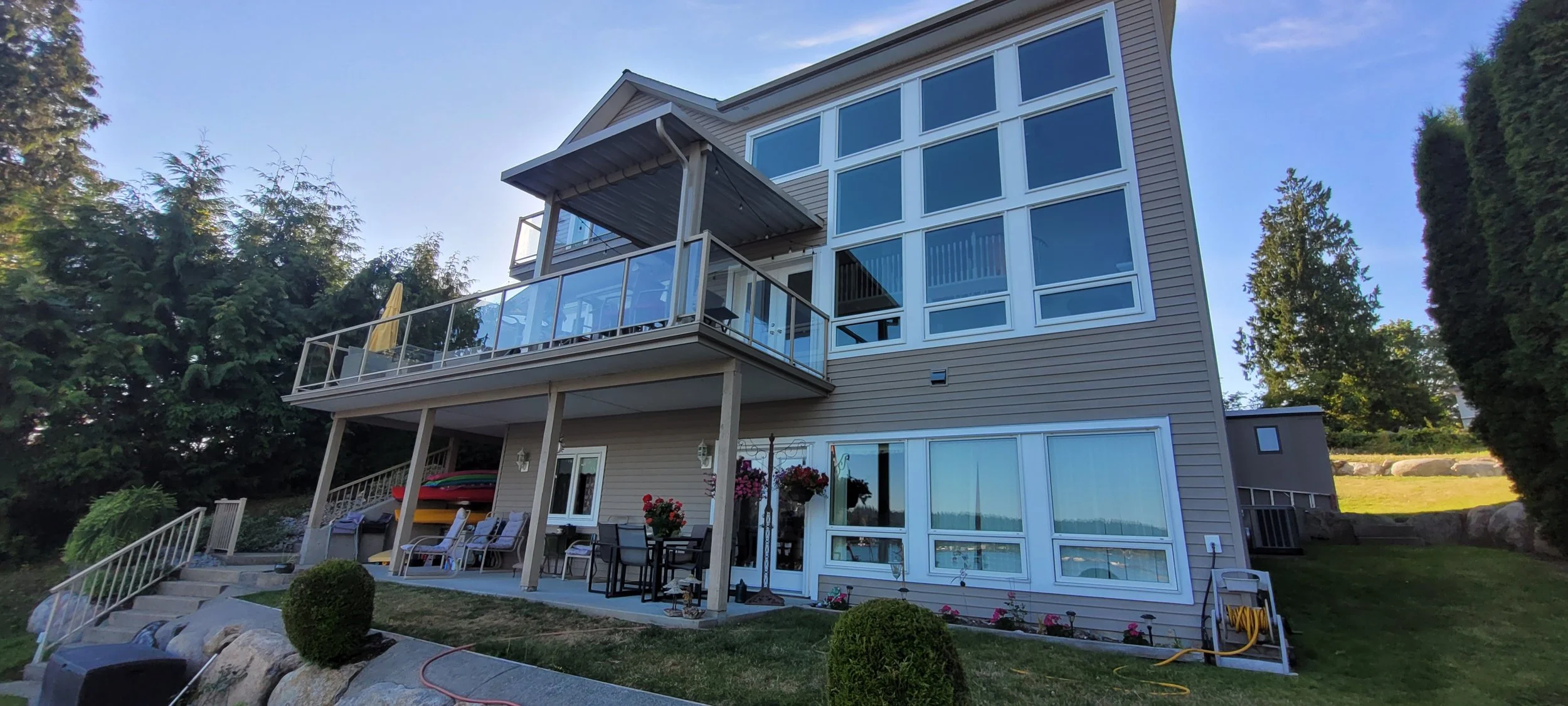 Multi-story house with large windows, a balcony, and a patio area with outdoor furniture and flowers, surrounded by trees.