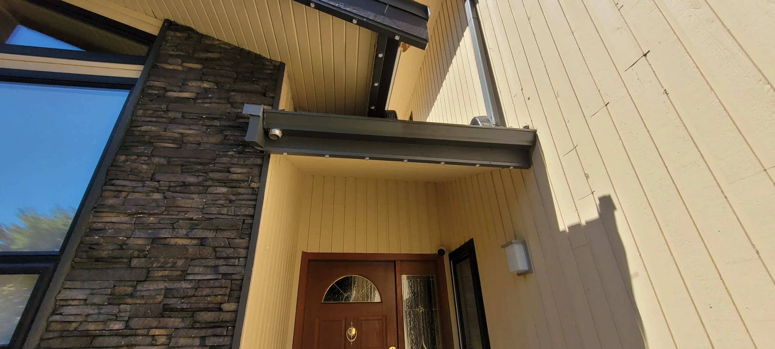 Front porch with yellow wooden siding, a wooden door with a semi-circular window, a black doormat, a black glass panel to the right of the door, a wall-mounted light fixture, and a metal support beam. The porch has a wood deck floor and a stone wall 