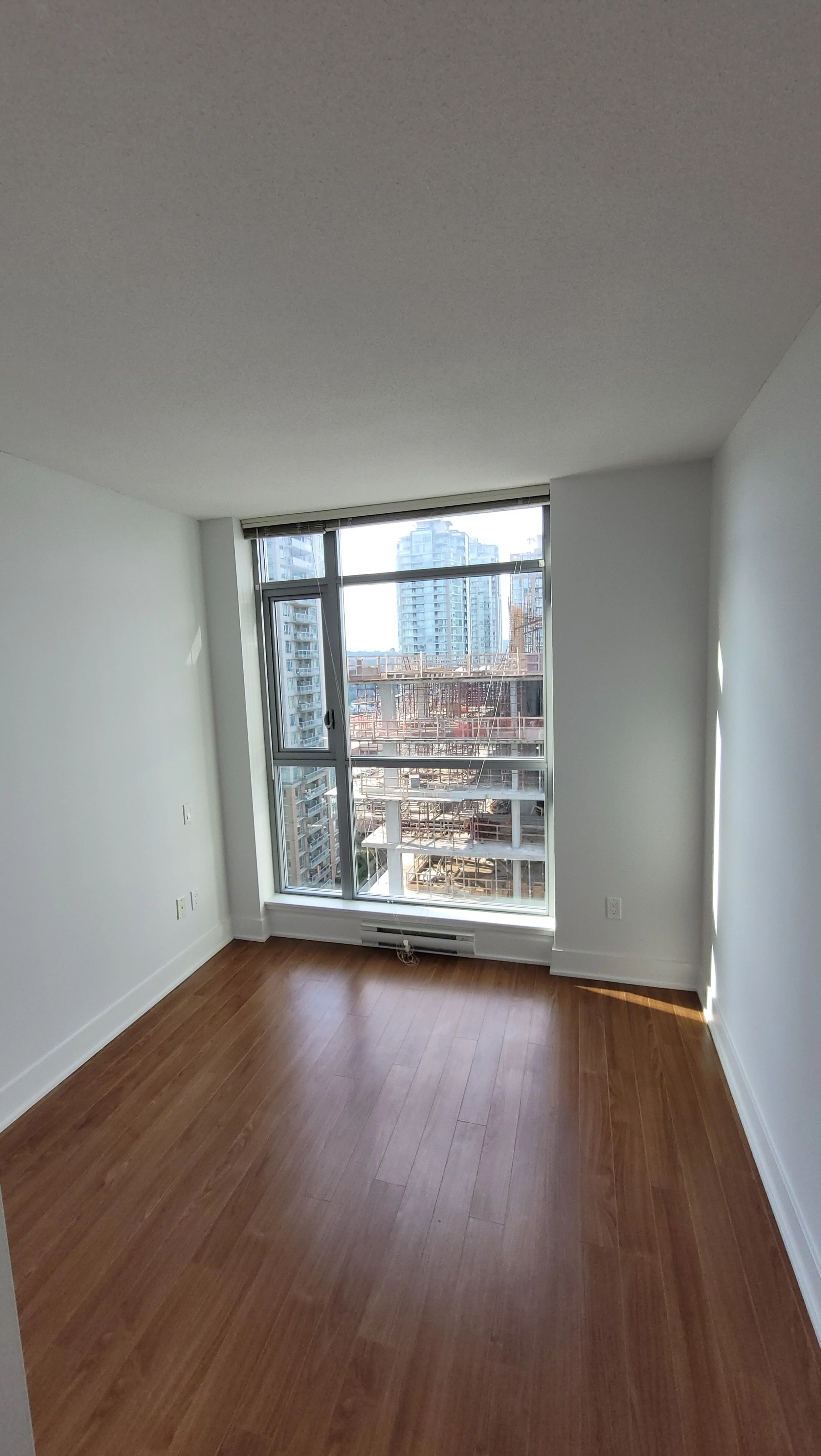 Empty room with large window, white walls, wooden floor, and cityscape view of buildings under construction.