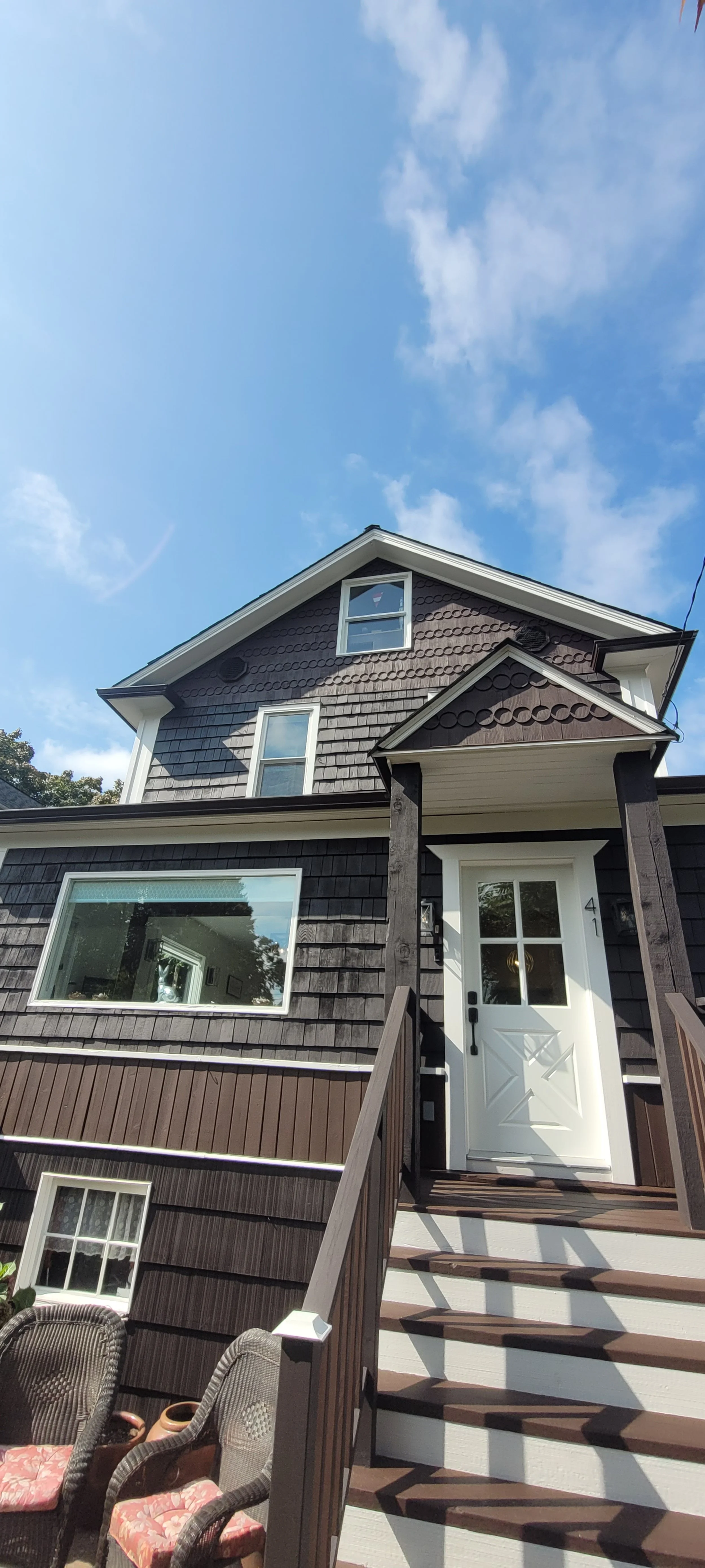 A two-story house with dark brown shingles, white trim, and a small front porch with stairs. A large window near the porch shows a reflection of trees. Outdoor chairs and potted plants are at the bottom of the stairs. The sky is clear with some cloud