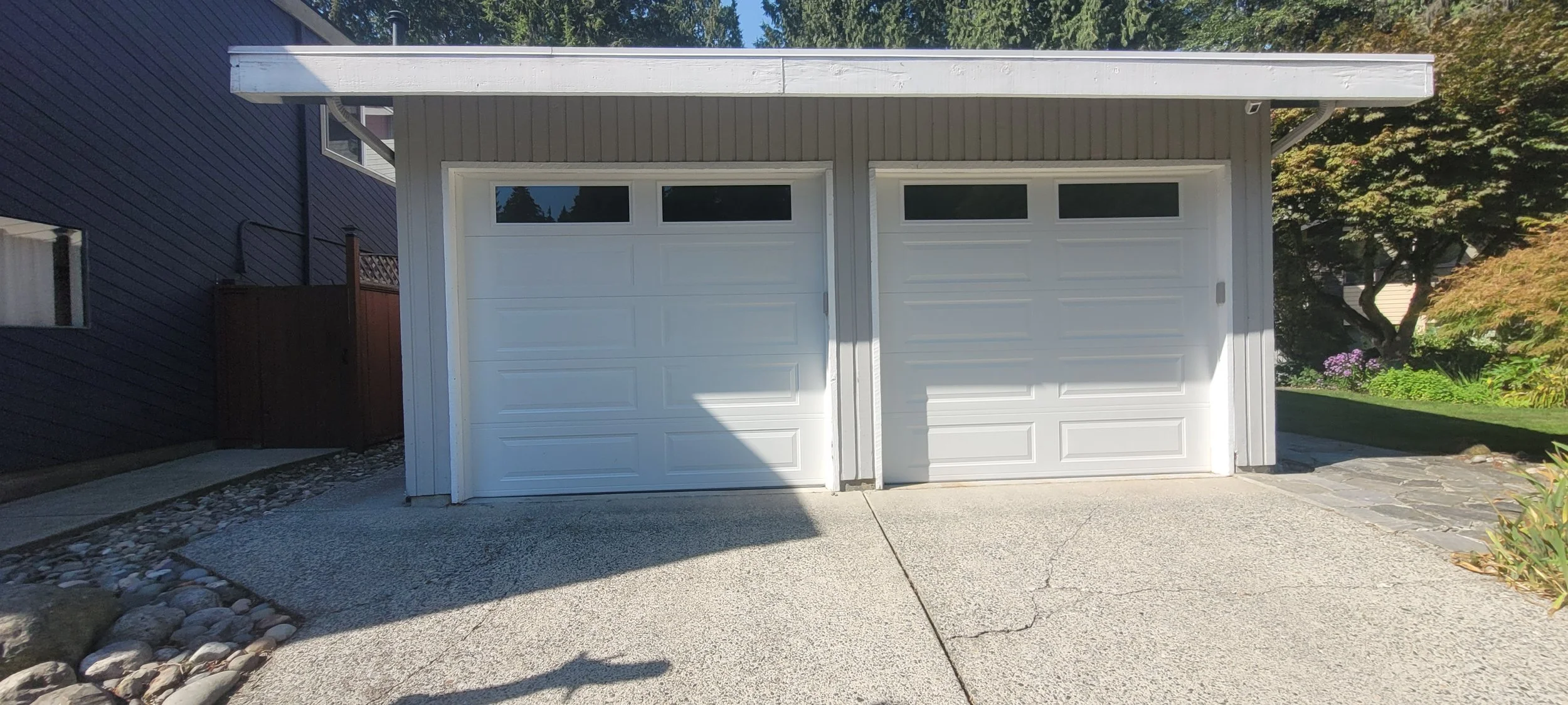 Two white garage doors side by side on a residential building, with a concrete driveway in front and a landscaped garden to the right, partially shaded by a tree.