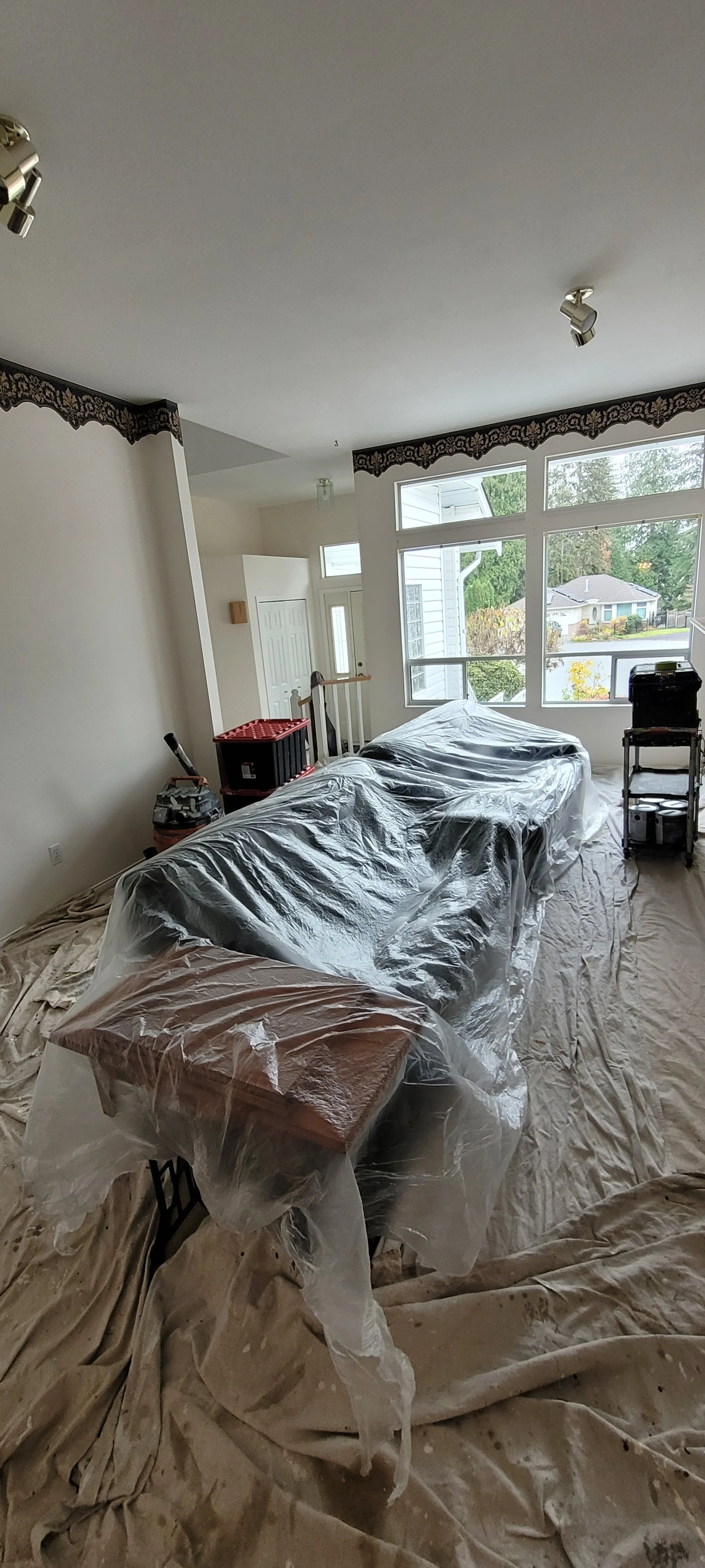 Living room with plastic-covered furniture and a tarp on the floor, indicating painting or renovation work in progress, near windows showing a suburban neighborhood.